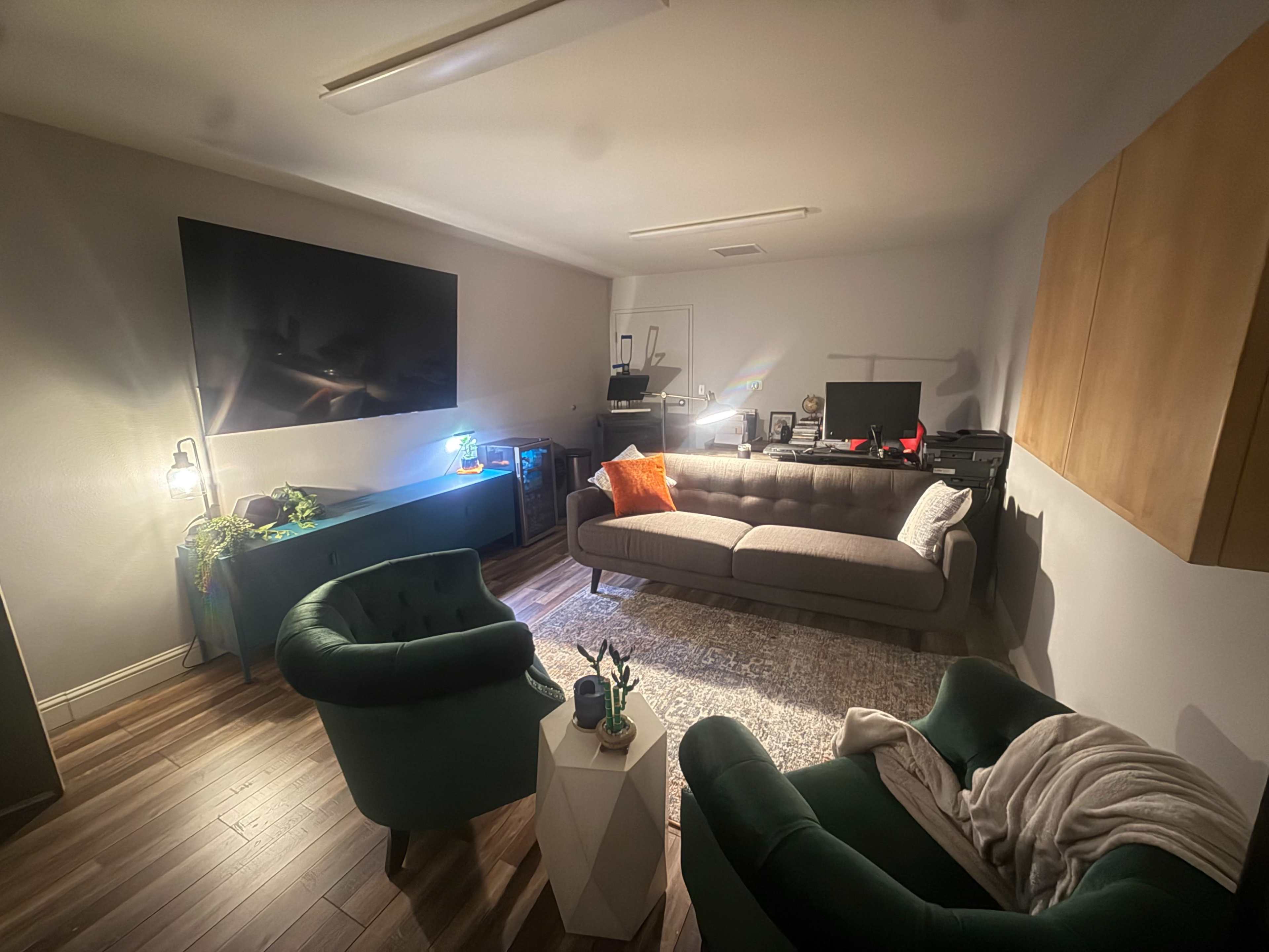 The image shows a cozy living room featuring a gray sofa, two green chairs, a coffee table, and a large wall-mounted TV.