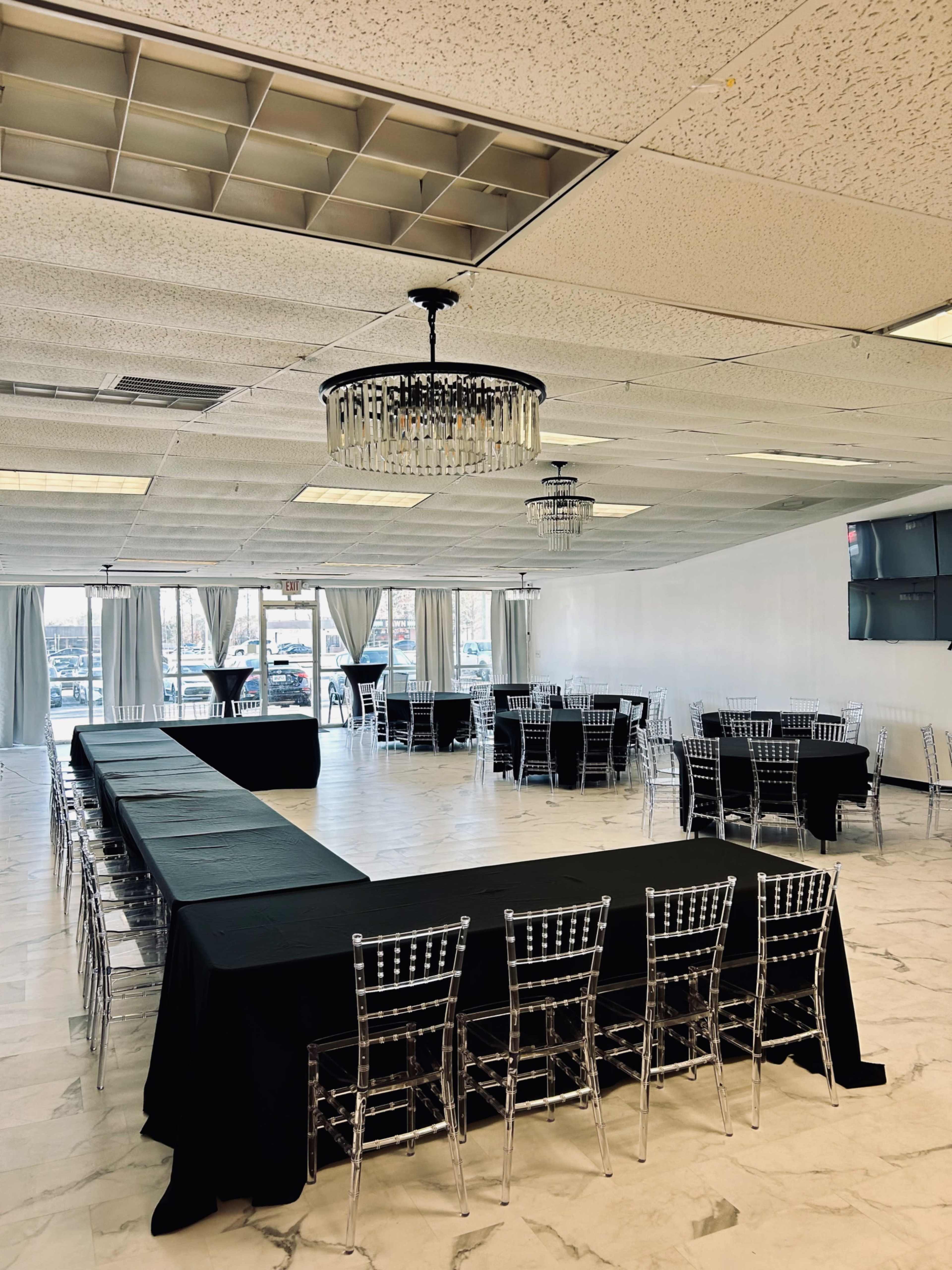 A spacious event room features rows of tables covered in black cloth, surrounded by transparent chairs, with chandeliers hanging from the ceiling.