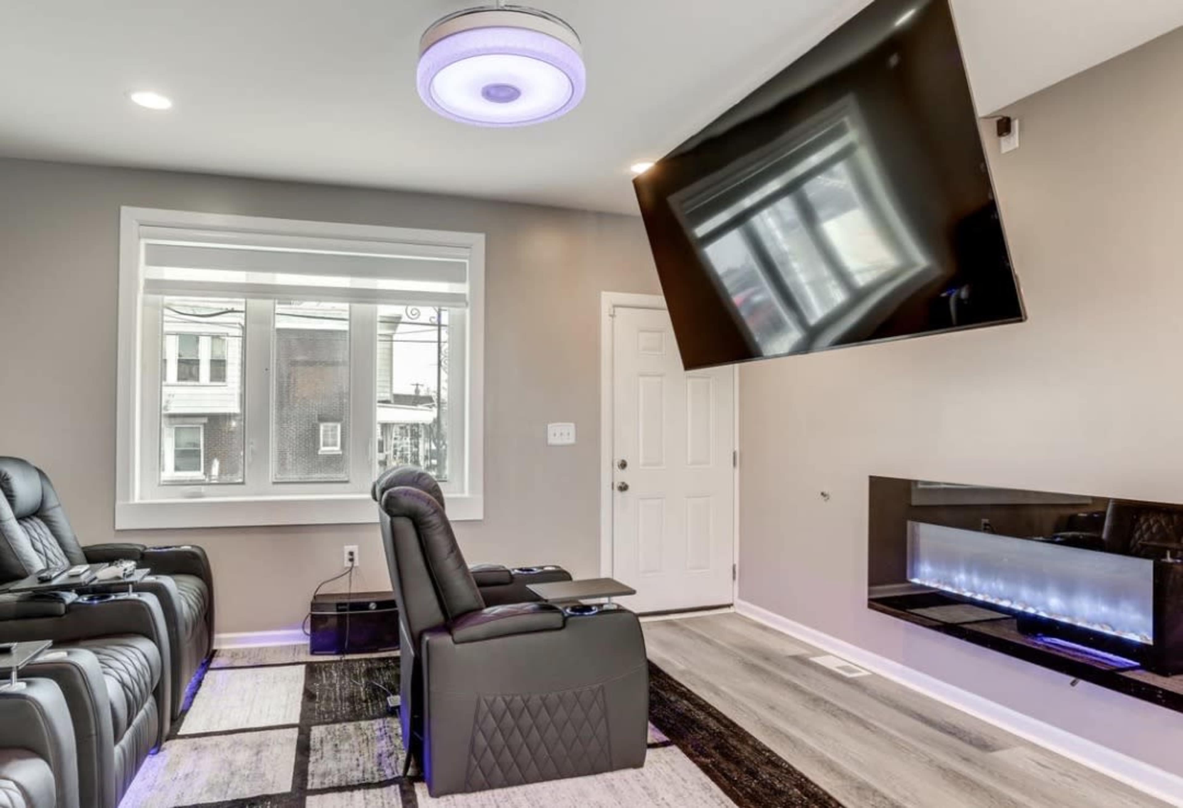 The image depicts a modern living room featuring two reclining chairs, a mounted television, and an electric fireplace under a large window.