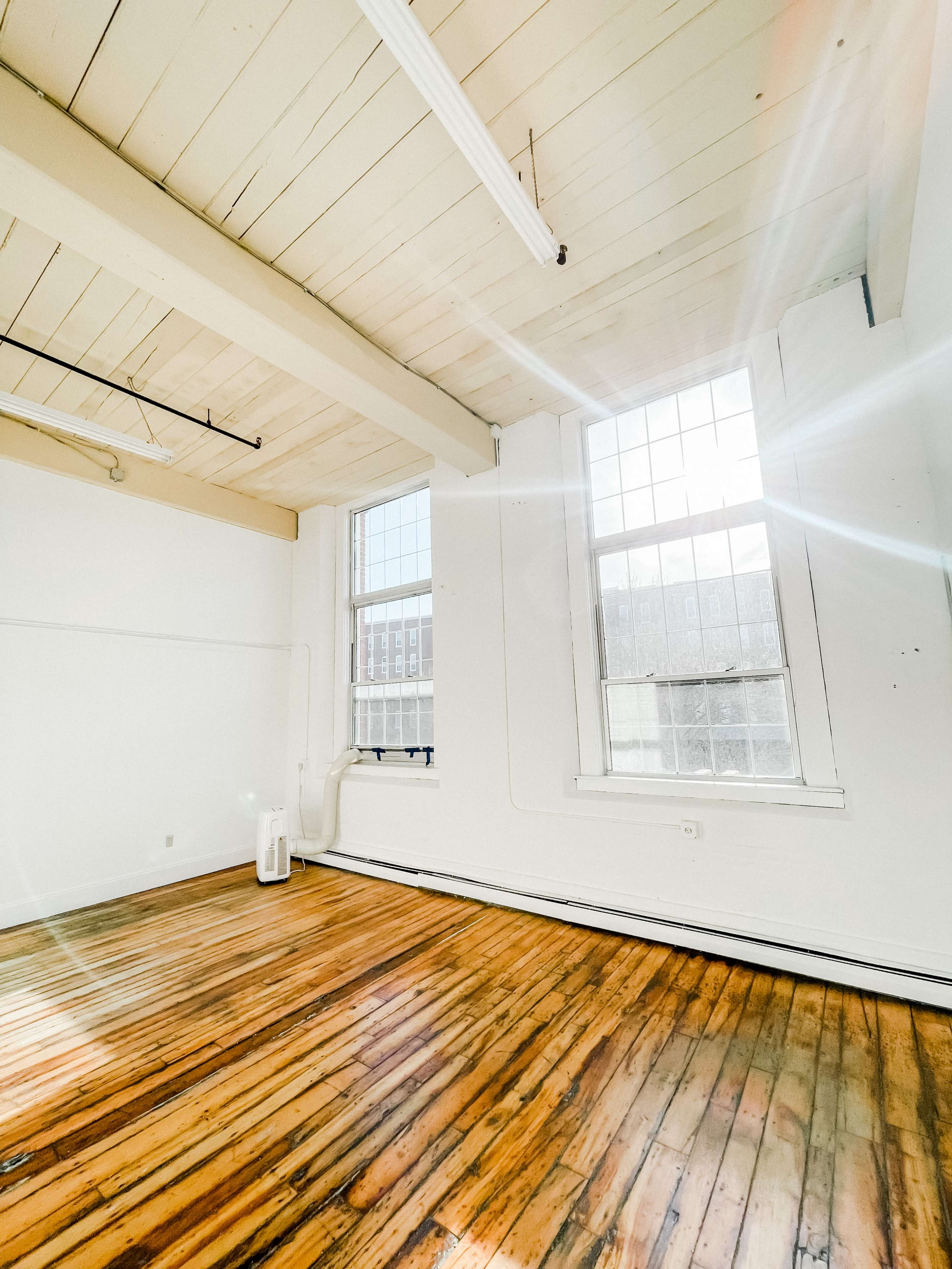 An empty room with large windows allowing natural light to illuminate the polished wooden floor and white walls.