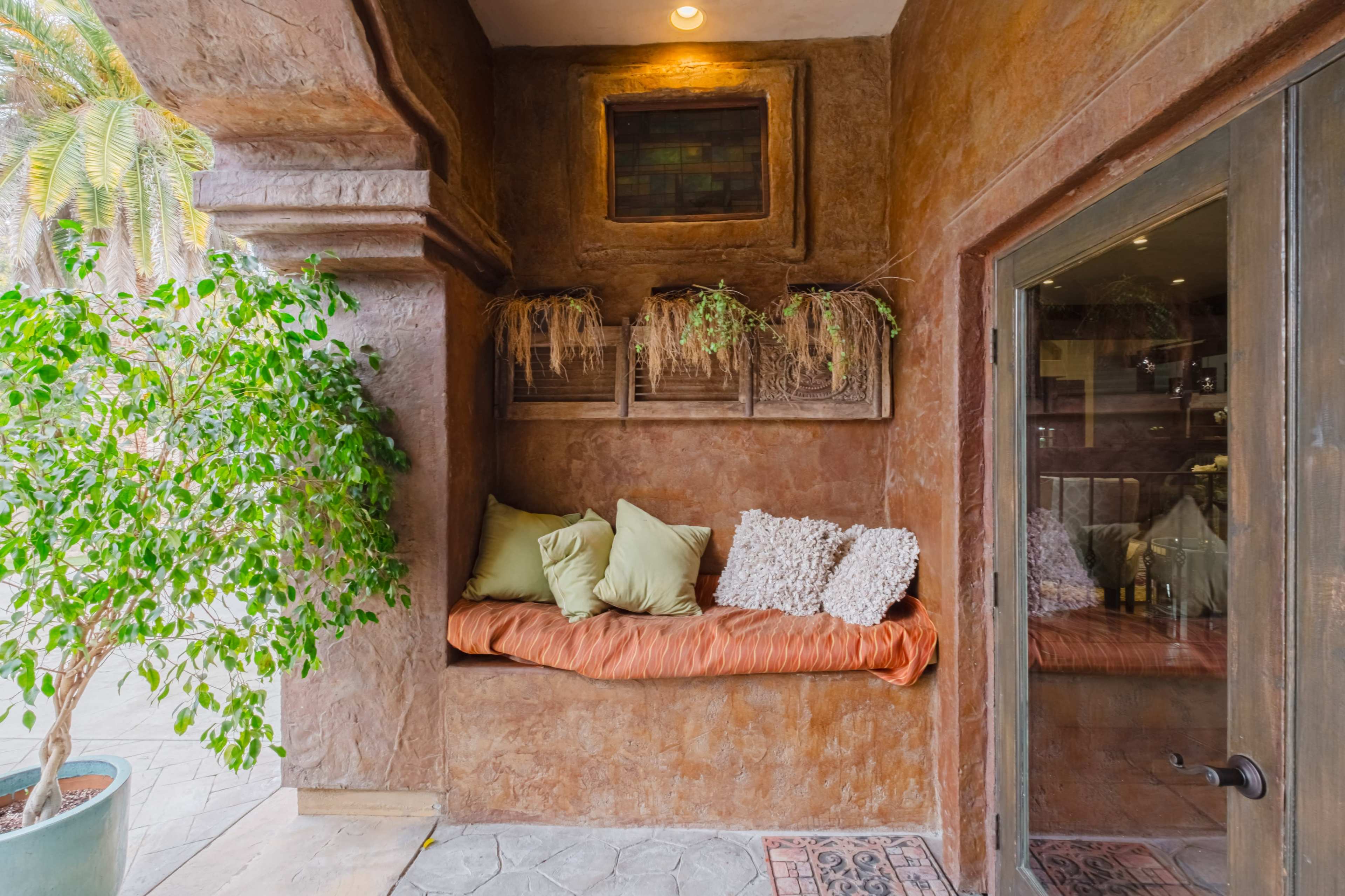 A seating nook is framed by textured walls and features green pillows on a copper-hued bench, with plants in wall-mounted boxes and a door leading to an interior space.