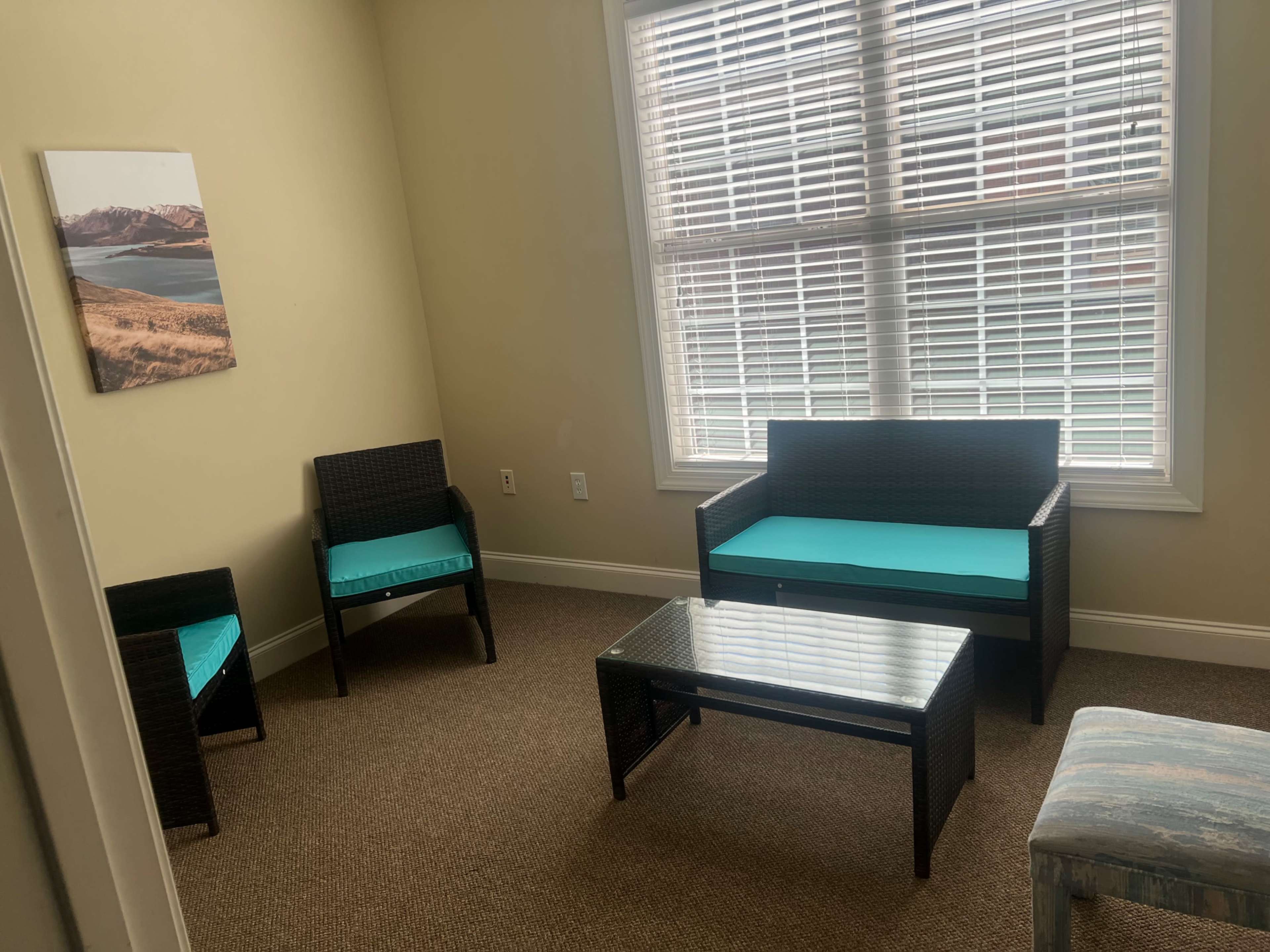 The image shows a small living area with two chairs and a sofa, all featuring turquoise cushions, accompanied by a glass coffee table, and a large window with blinds letting in natural light.