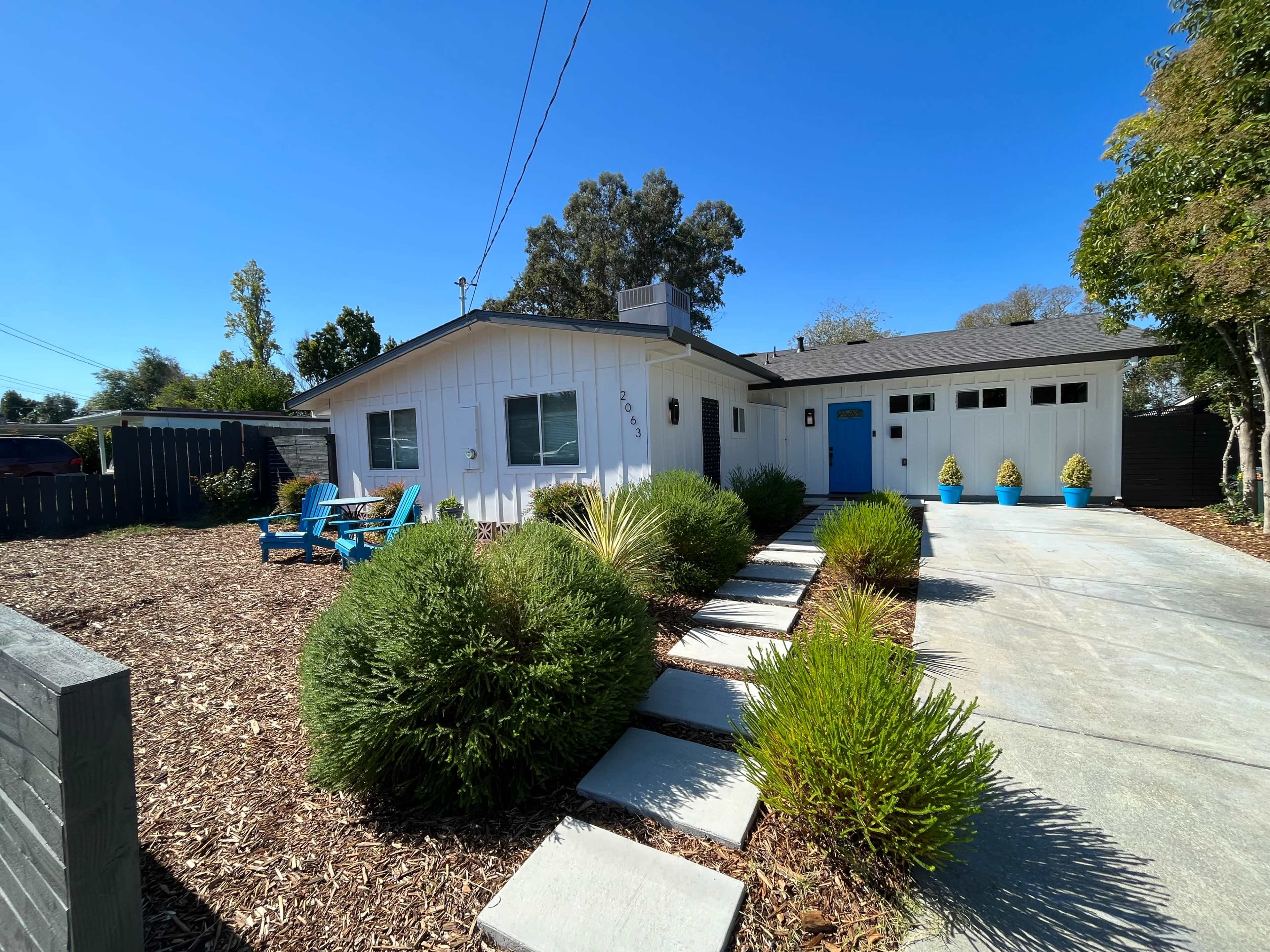 A single-story white house with a blue door is surrounded by stone walkways and landscaped shrubs in the front yard.