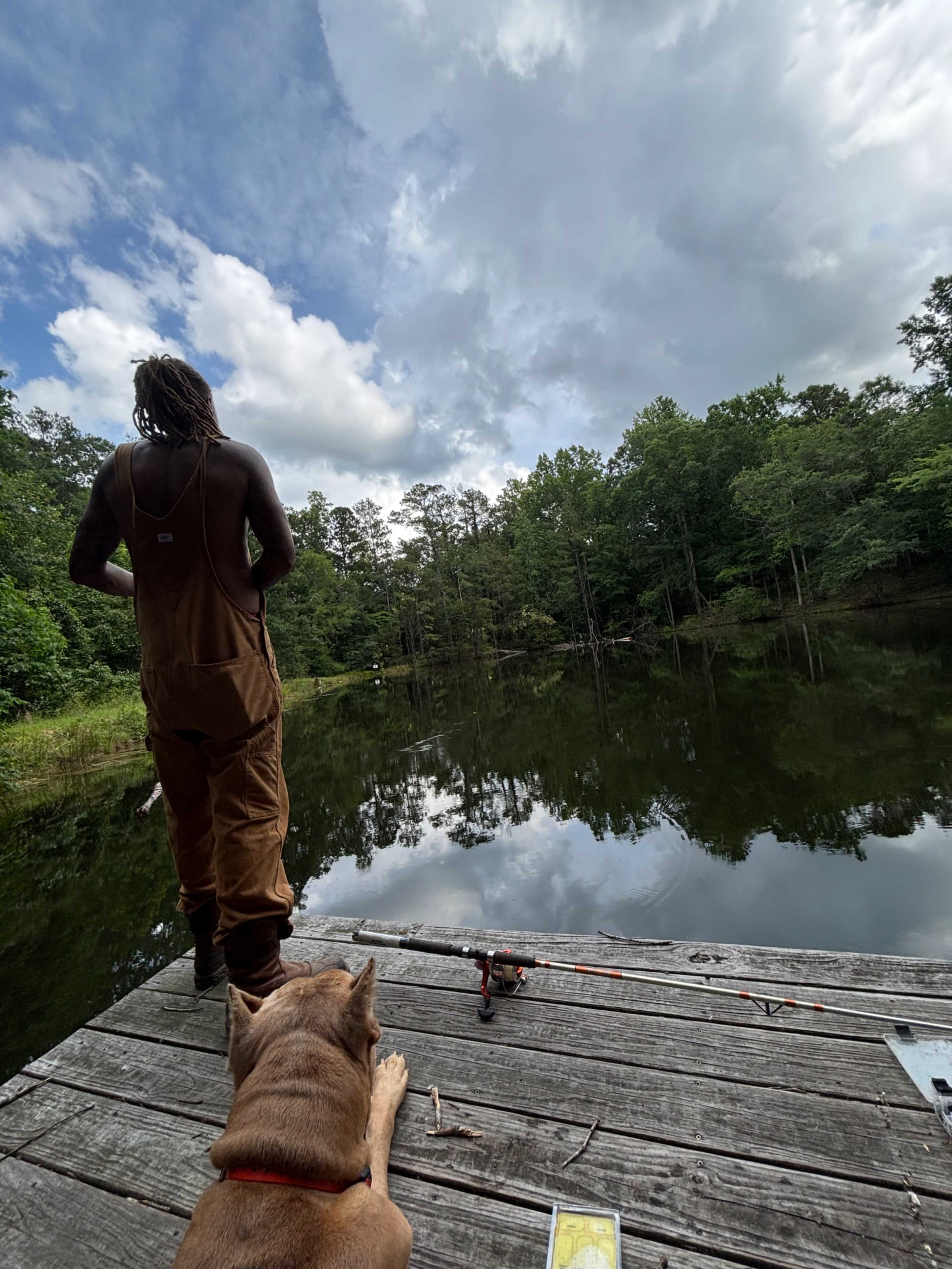 A person in overalls stands on a wooden dock overlooking a calm pond, while a dog rests nearby.