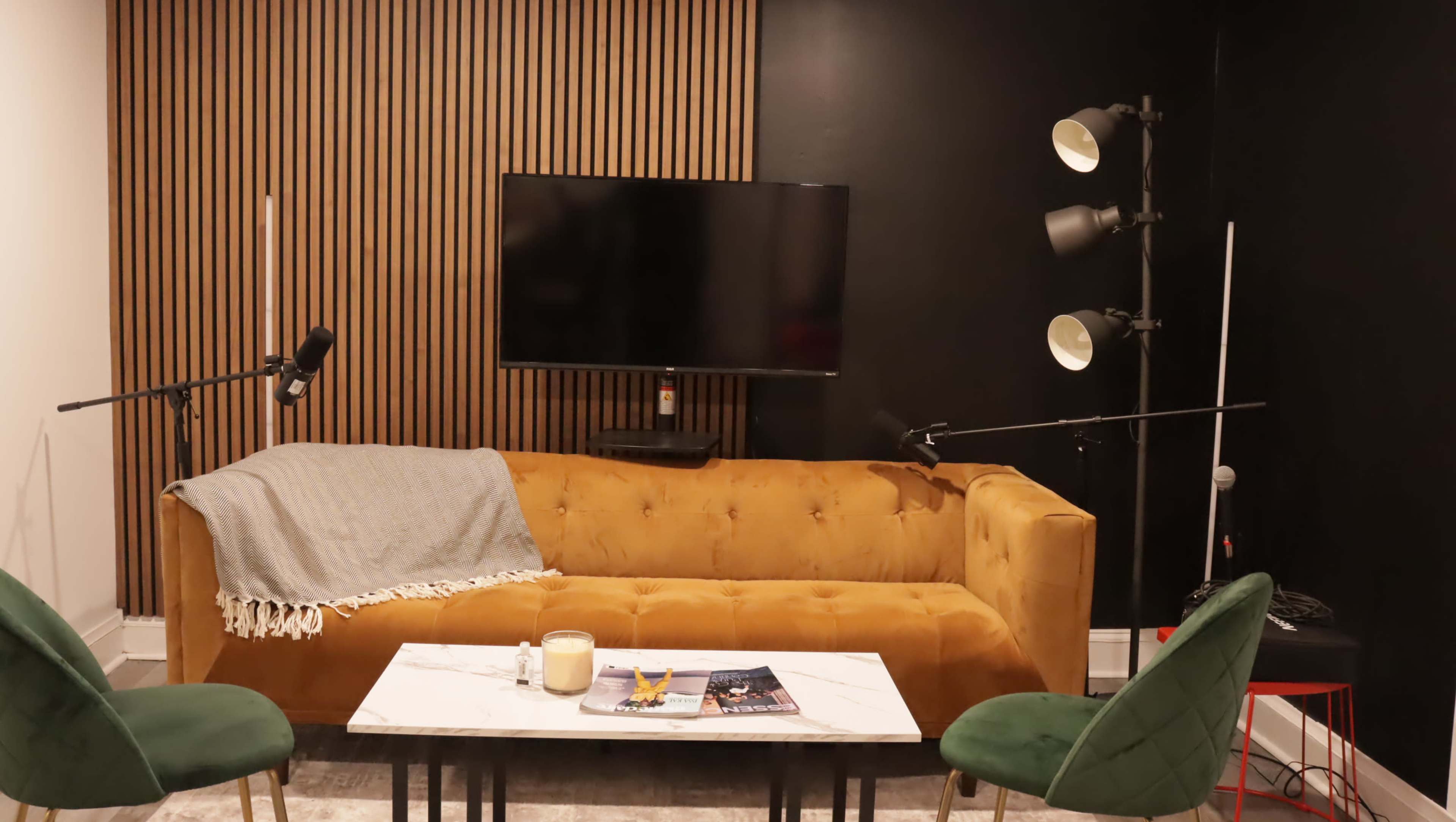 A cozy living room features a mustard-colored couch, a coffee table, a wall-mounted TV, and two green chairs, with microphones set up for recording.