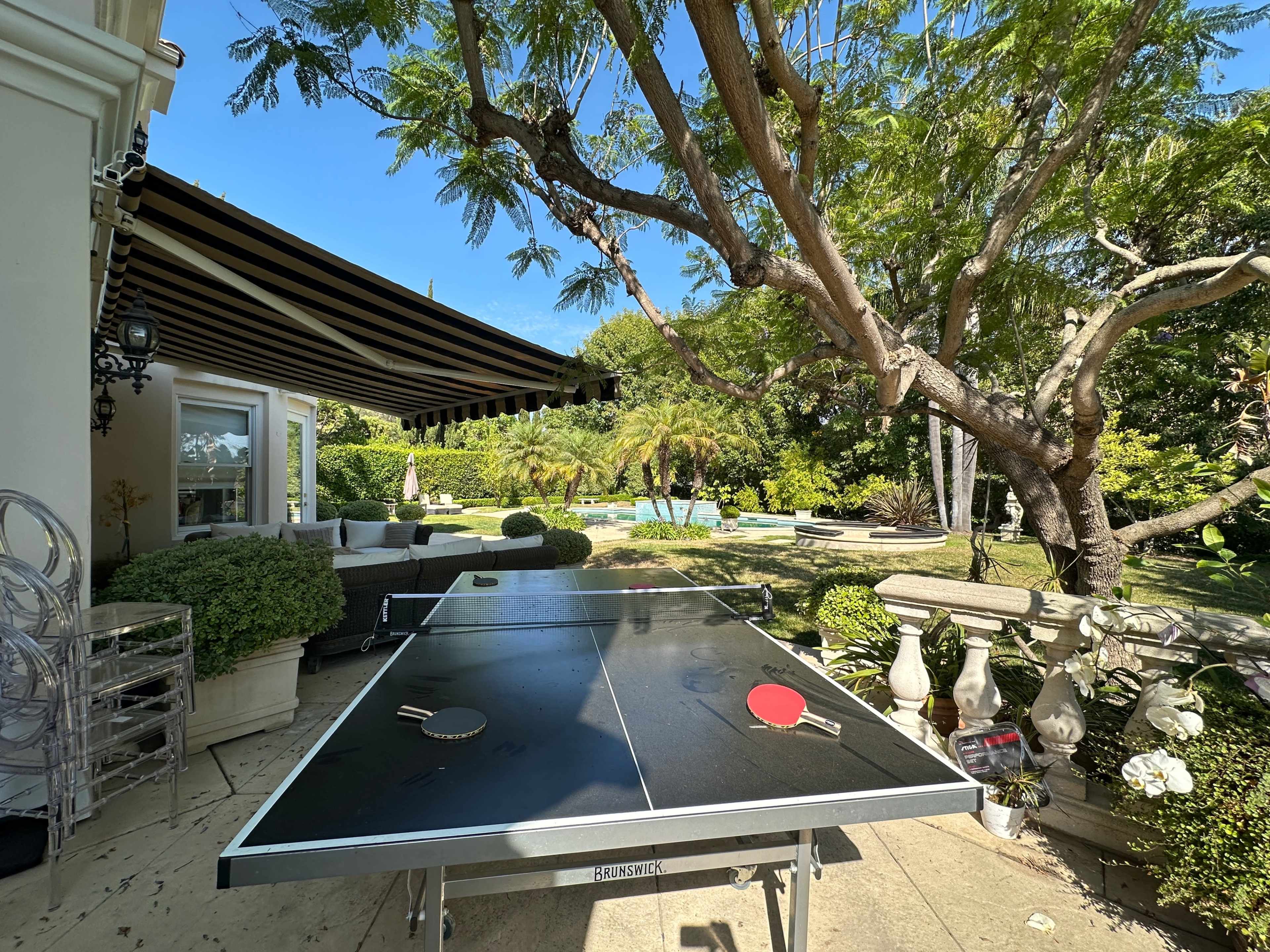 A ping pong table is set up in a garden with a large tree and a swimming pool in the background.