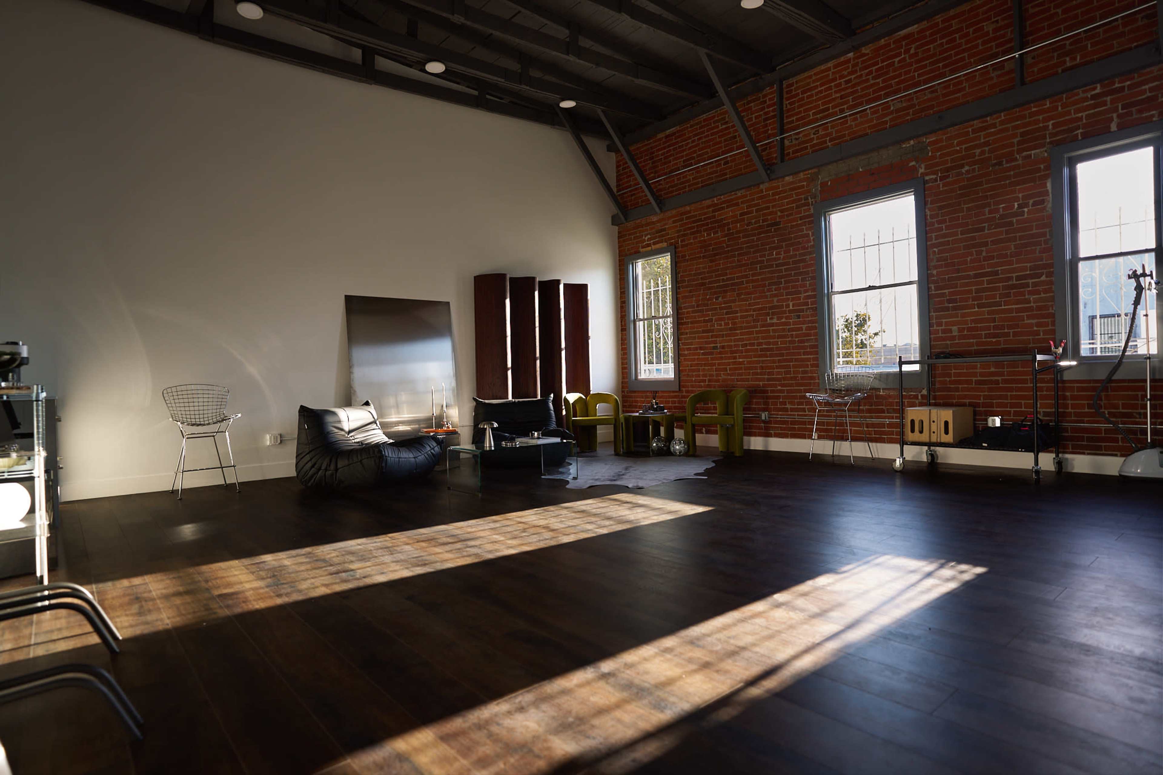 The image shows a spacious, well-lit room with exposed brick walls, wooden flooring, and modern furniture, including a bean bag and chairs.