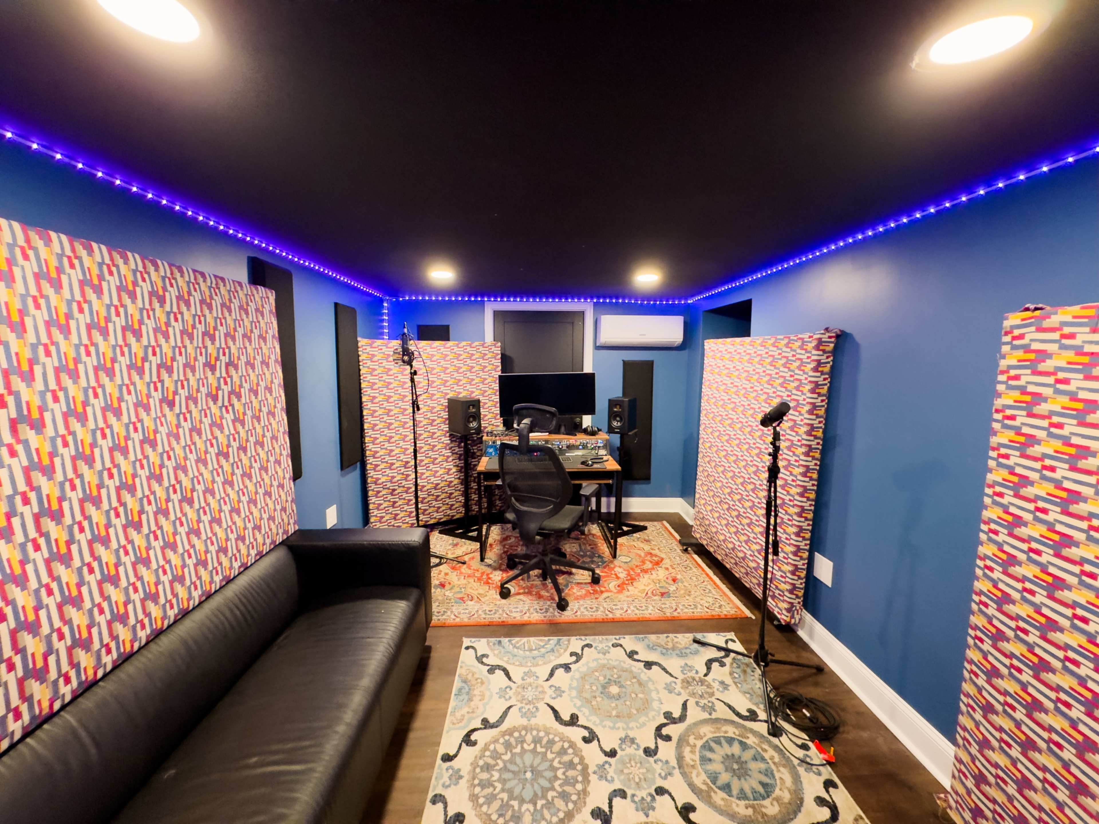 A music recording studio with soundproofing panels, microphones, and a desk equipped with audio equipment, illuminated by LED lights along the walls.