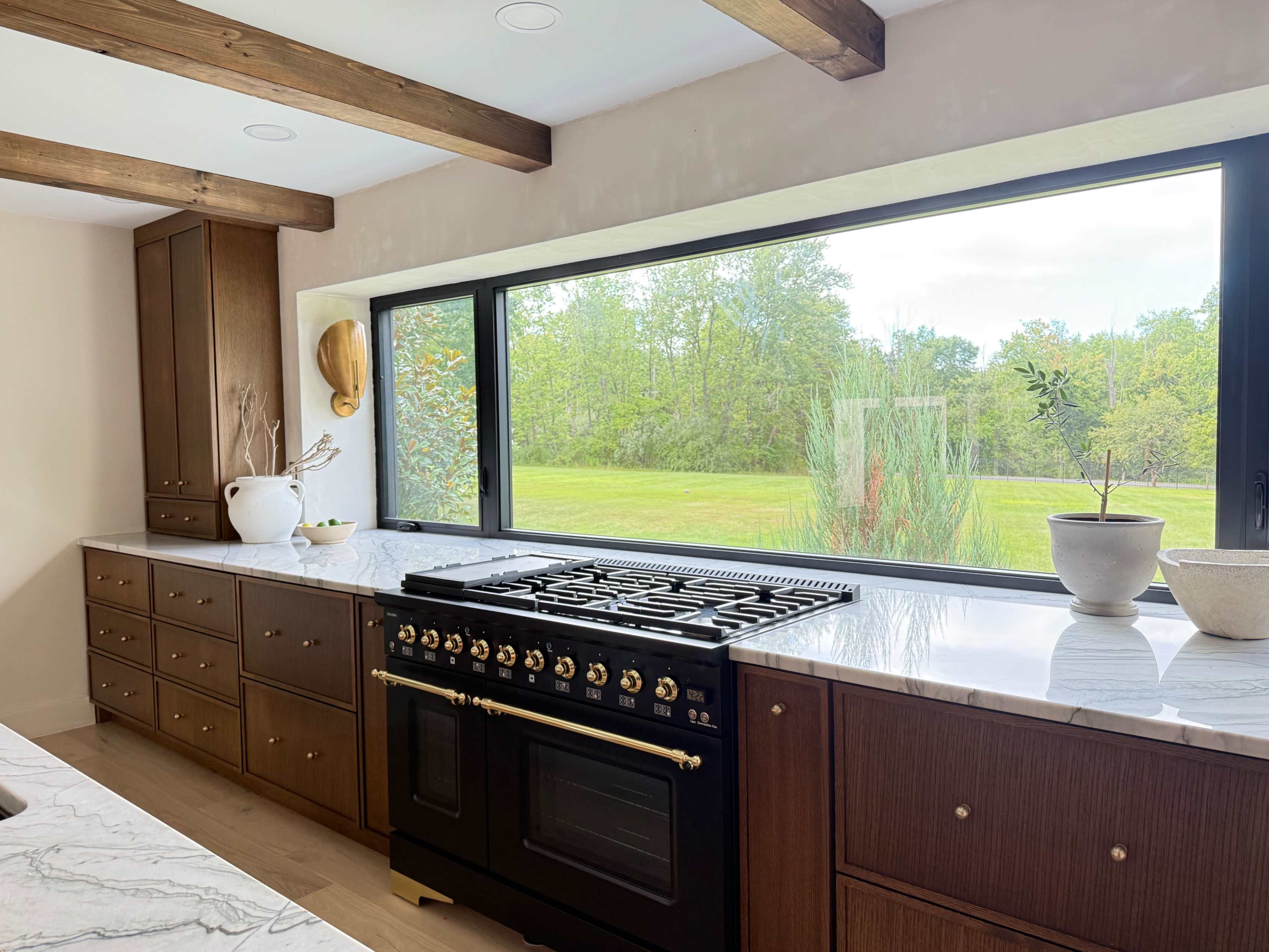 A kitchen features a black stove with a marble countertop and a large window overlooking a green landscape.