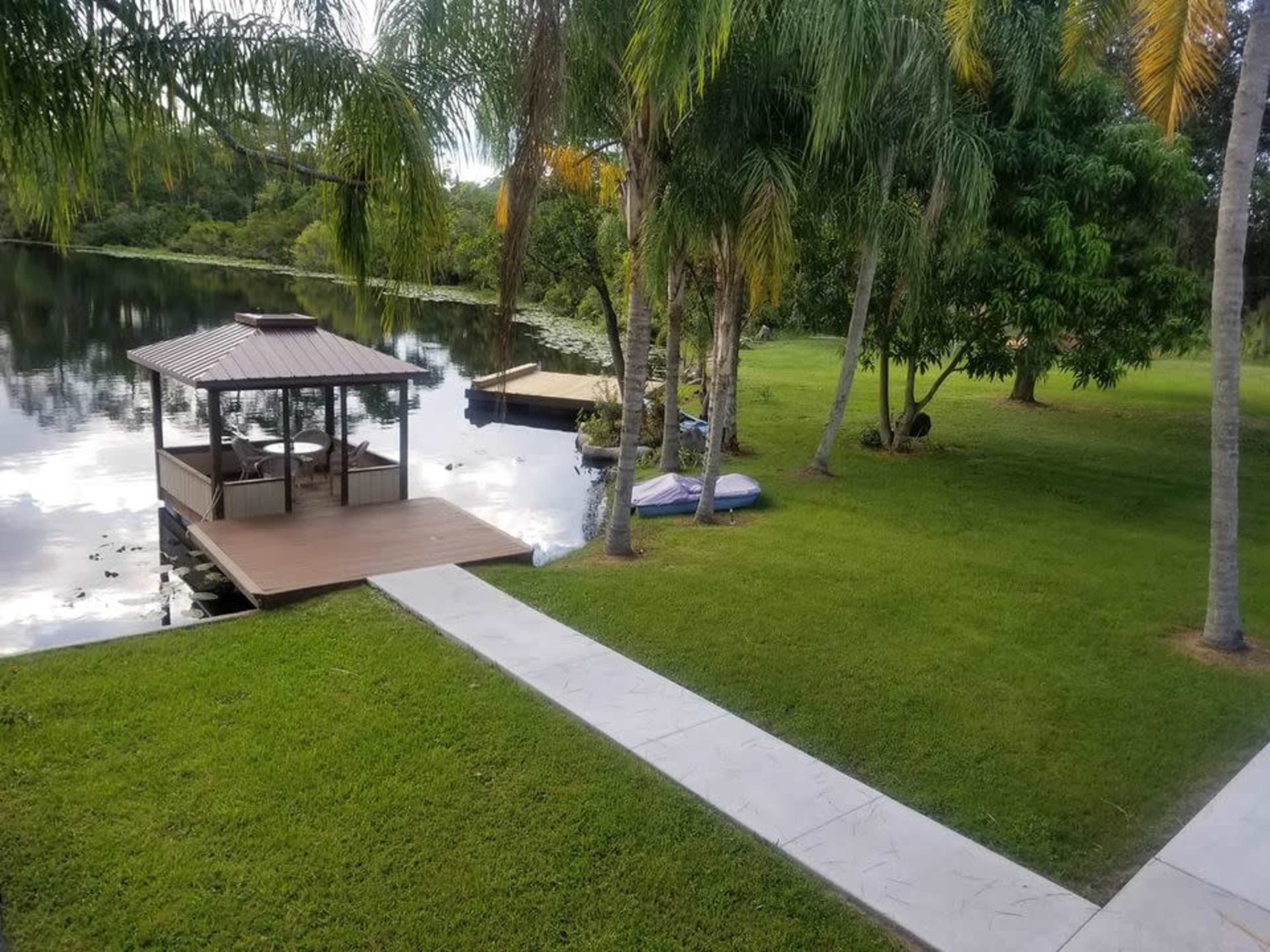 A serene lakeside area featuring a wooden dock with a gazebo, surrounded by green grass and palm trees.
