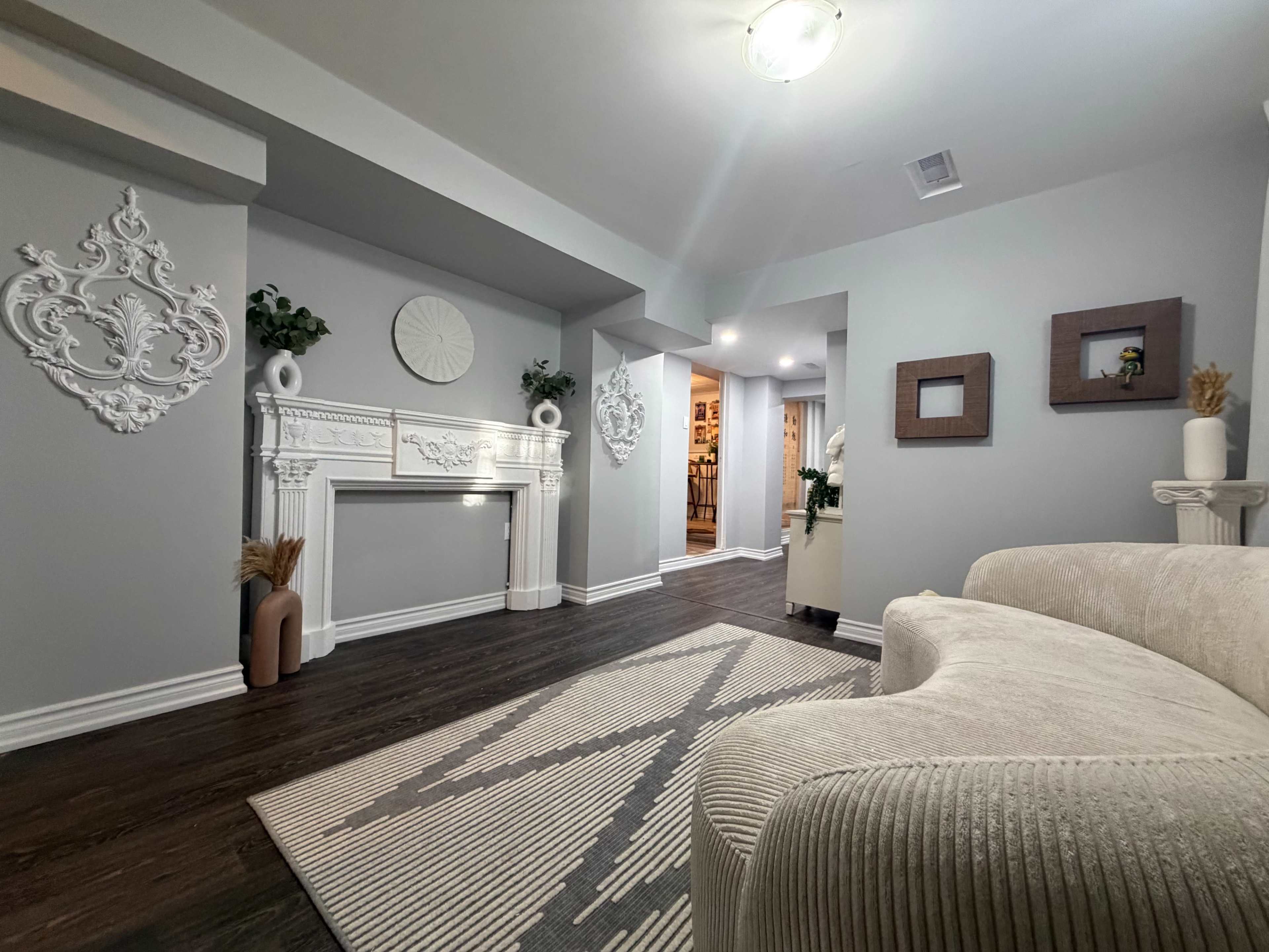 The image shows a modern living space with a light gray color scheme, featuring a decorative fireplace, abstract wall art, and a plush sofa placed on a geometric-patterned rug.
