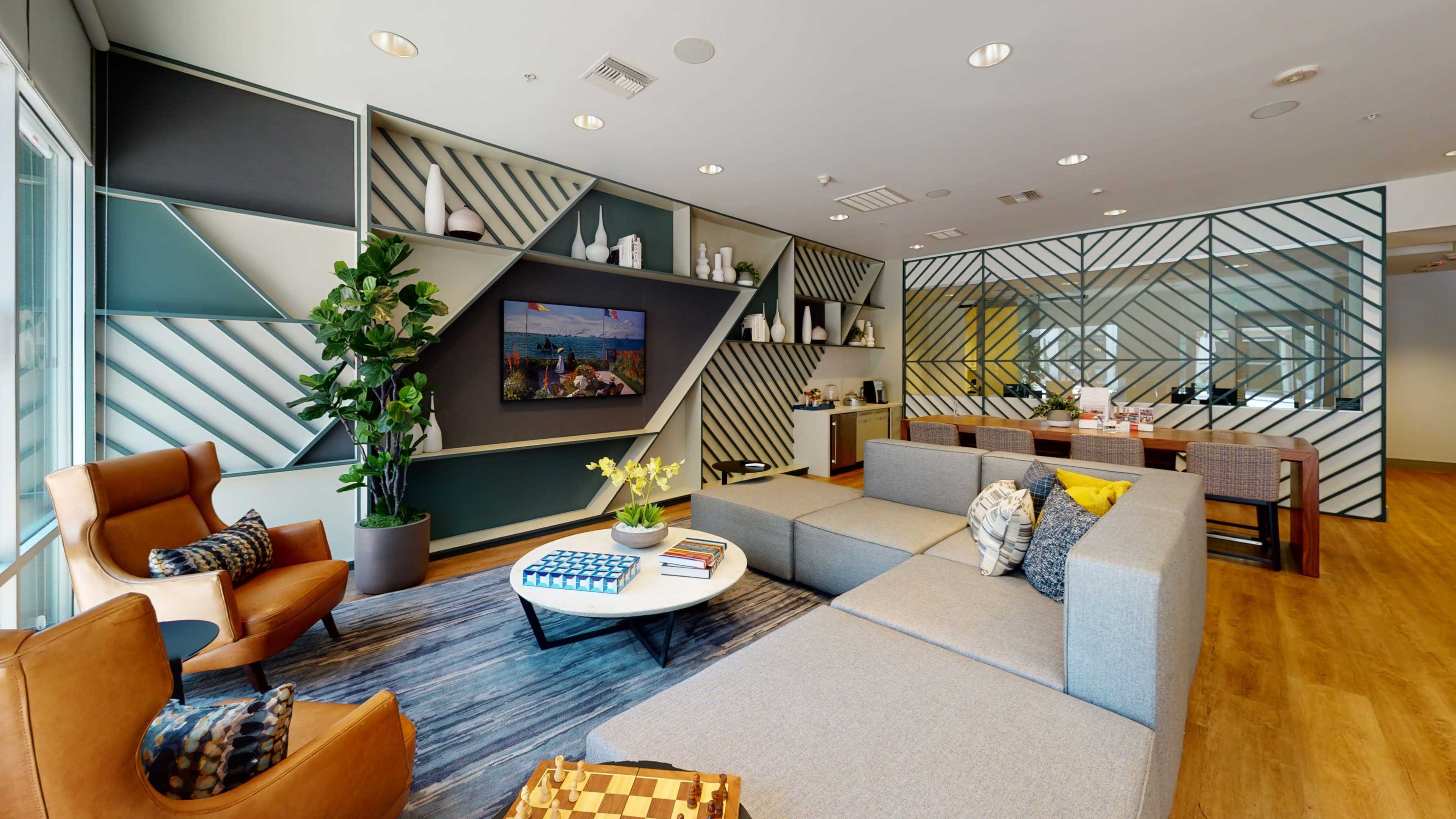 A modern lounge area featuring a sectional sofa, a coffee table, a television on a wall, and a dining table with chairs, surrounded by geometric wall decorations and indoor plants.