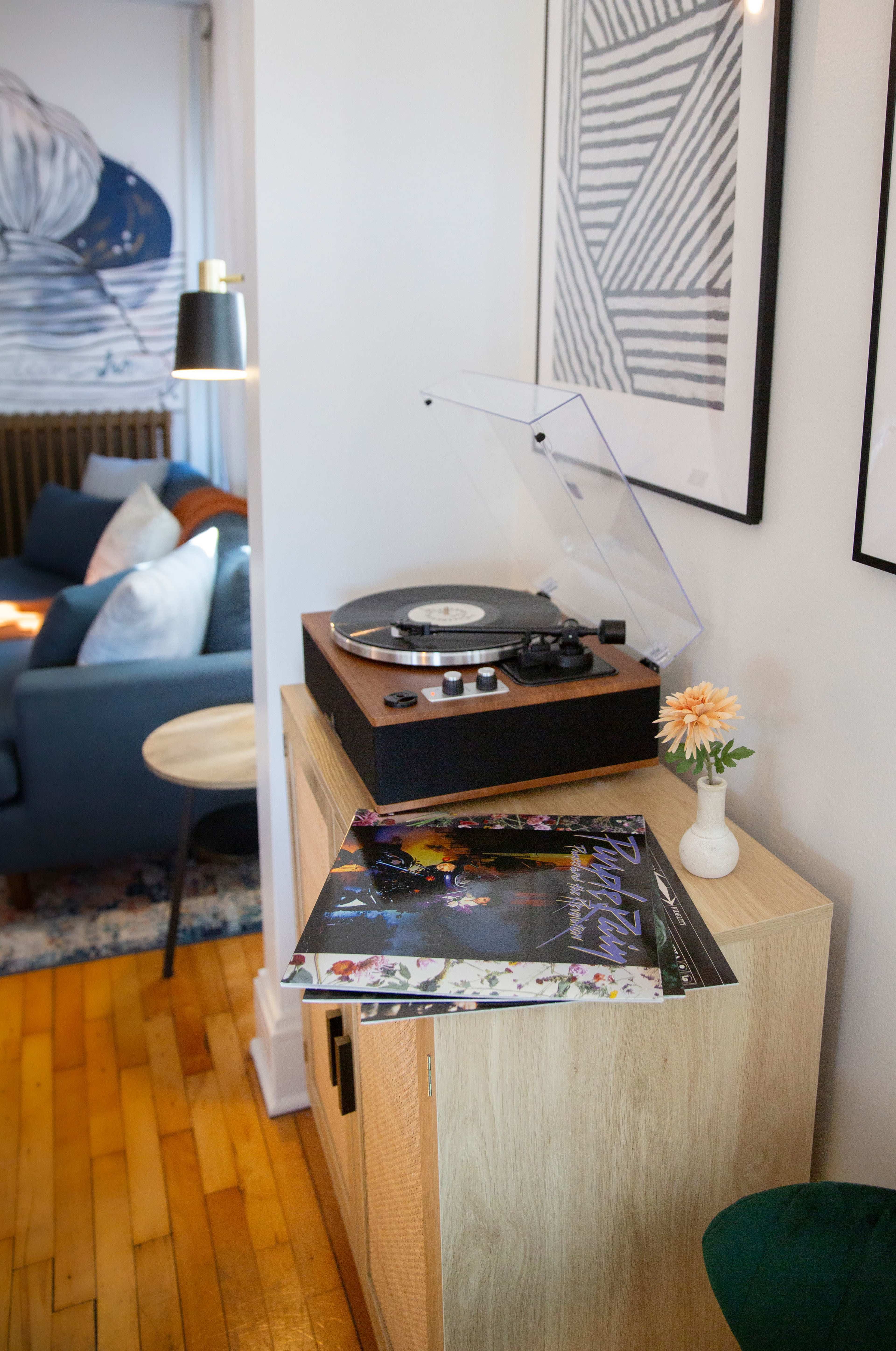 A wooden record player sits on a cabinet next to an album cover in a room with artwork on the walls and a couch in the background.