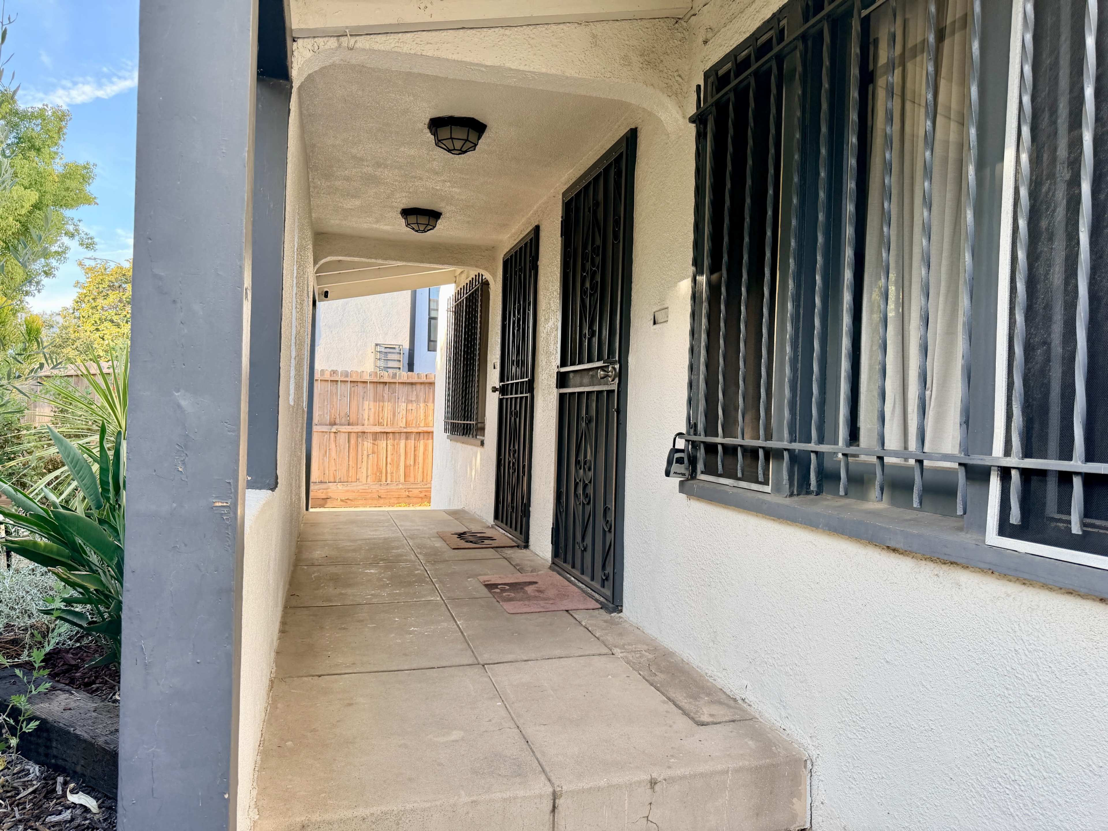 The image shows a covered porch area with two doors featuring decorative metal security bars, flanked by light fixtures and bordered by a garden.