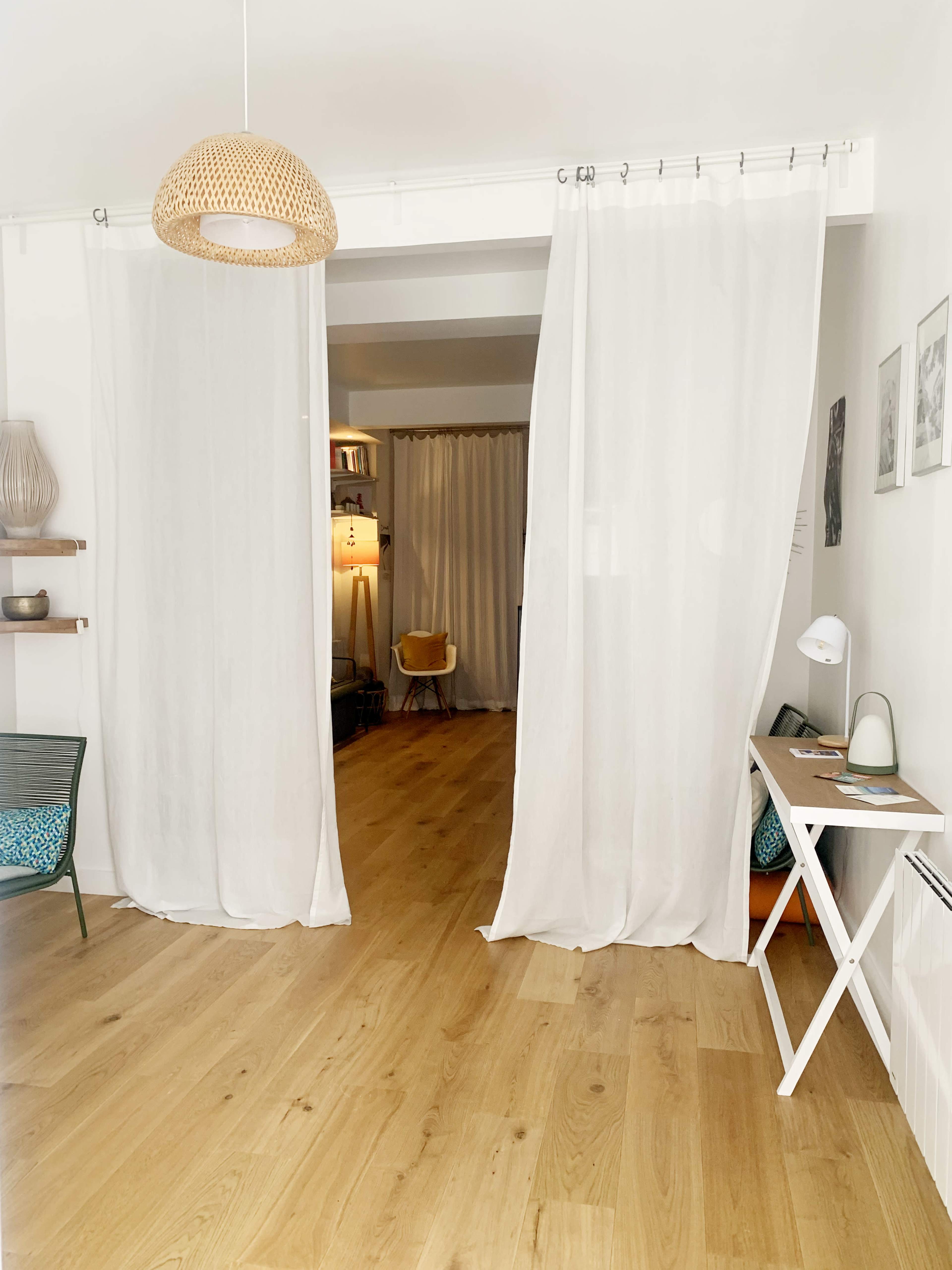 The image shows a bright interior space adorned with white sheer curtains separating two areas, with wooden flooring and minimalistic furniture.