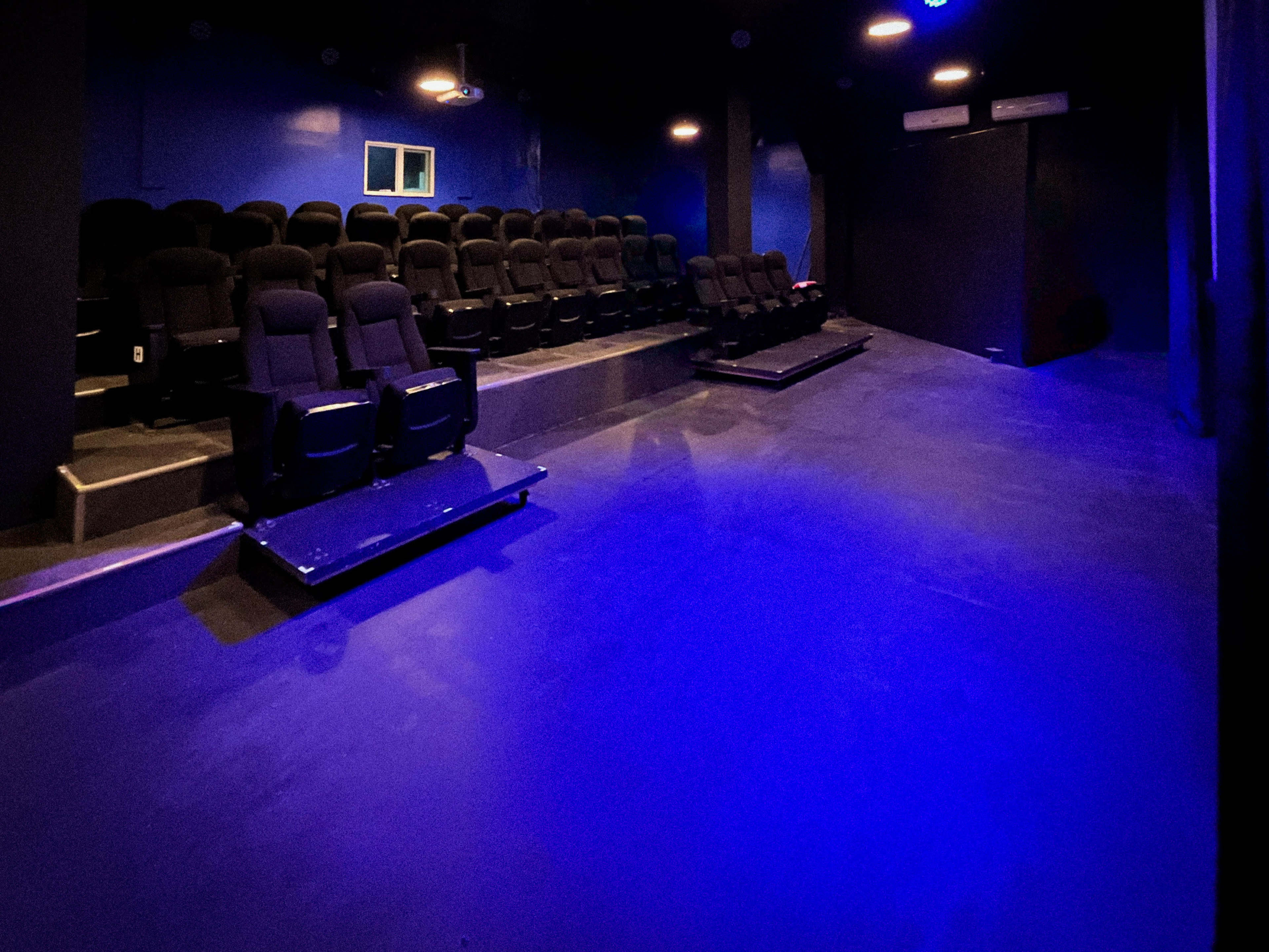The image shows a dimly lit theater with black seating arranged on risers and blue walls.