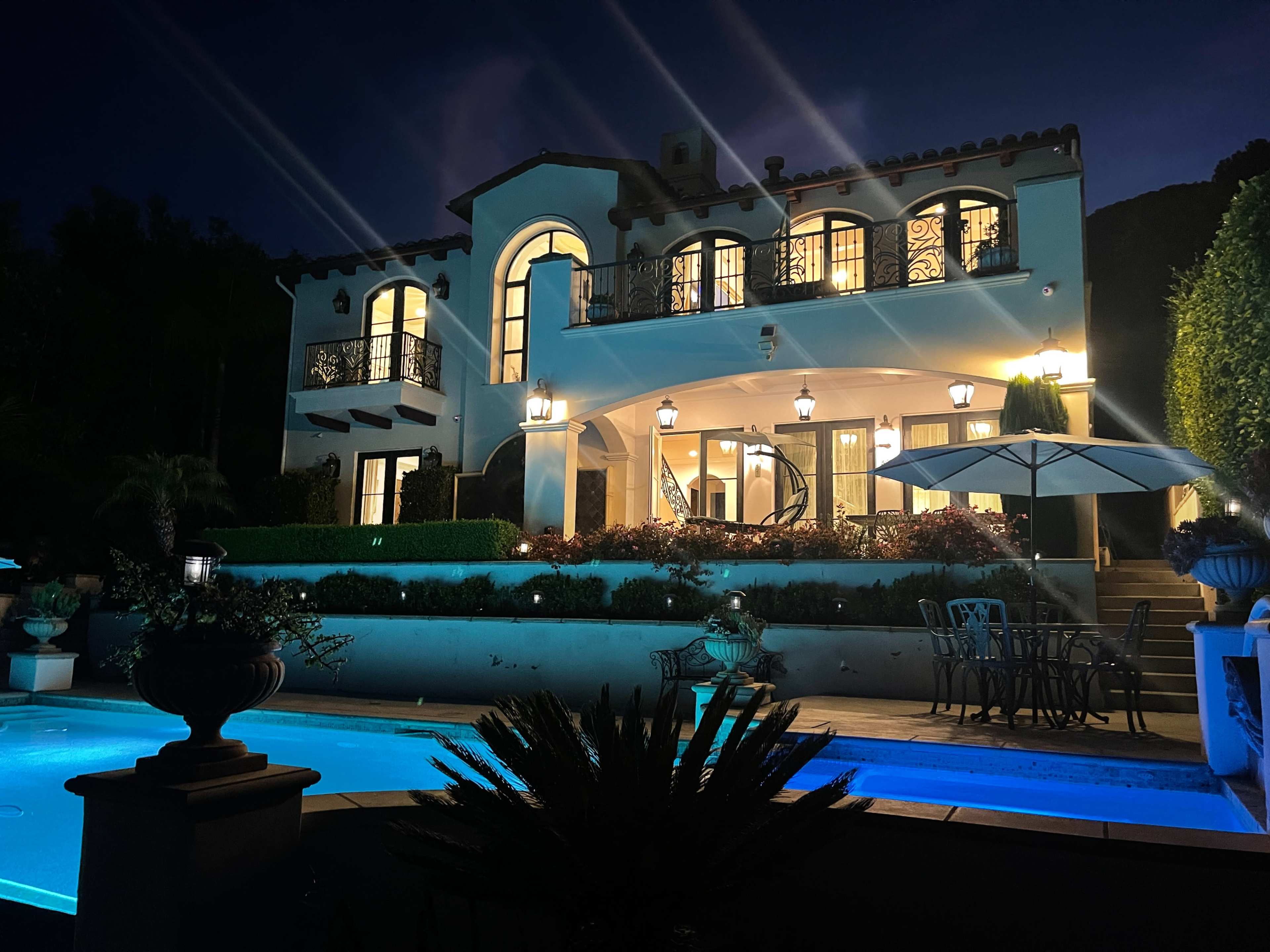 A large, illuminated villa with arched windows and decorative balconies overlooks a serene pool area at night.