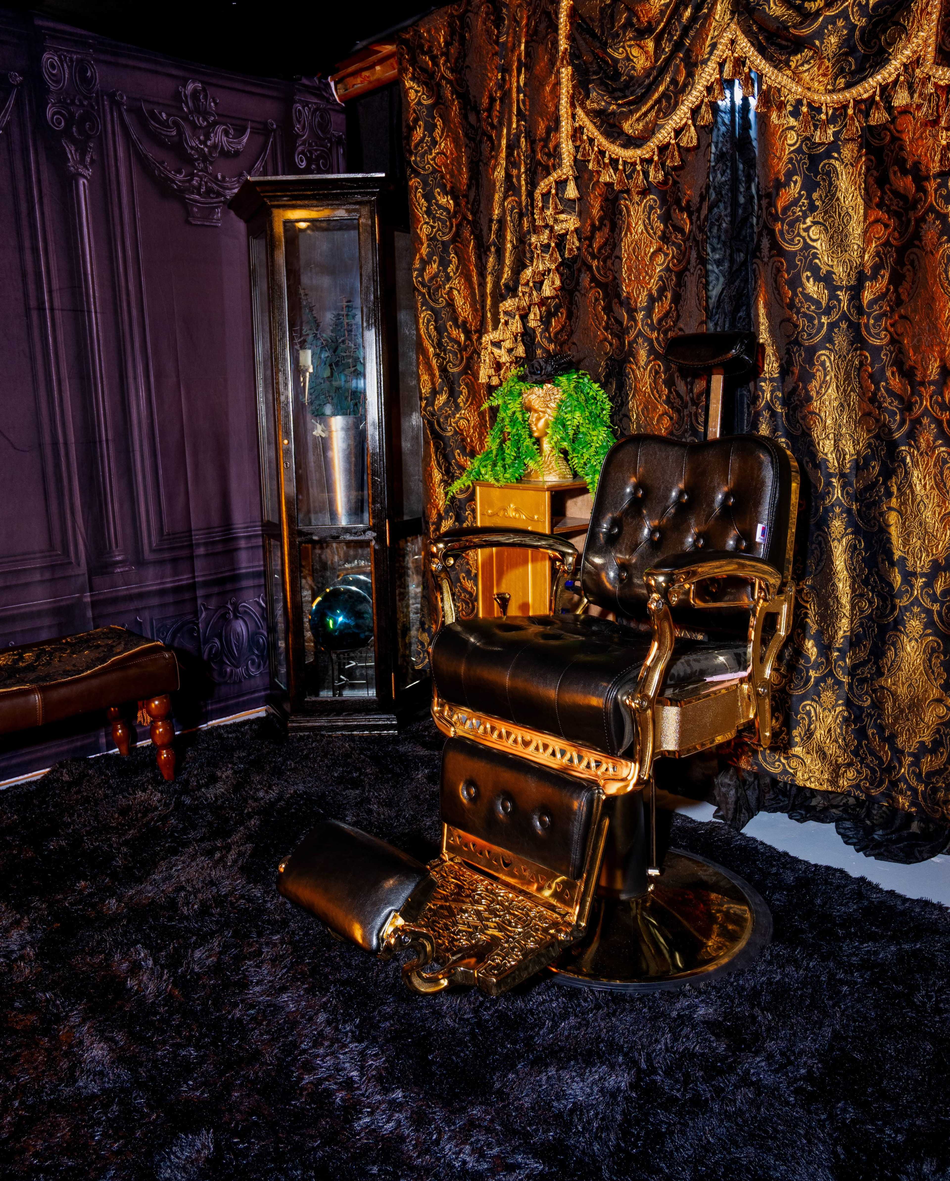 The image features an ornate barber chair made of black and gold, positioned on a dark, plush rug in a room adorned with rich, patterned drapes and decorative elements.