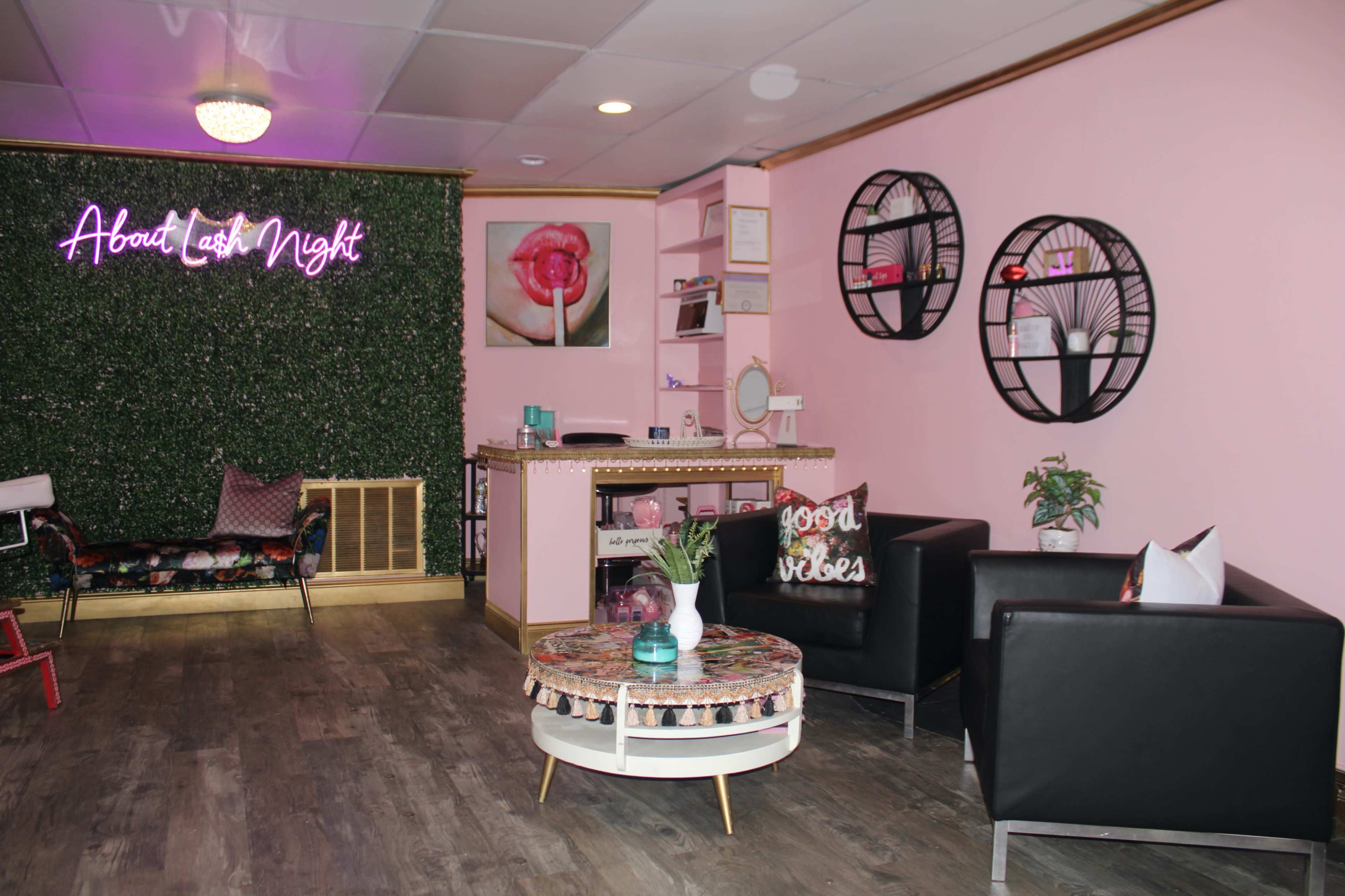 A cozy salon interior with pink walls, a green plant wall, a neon sign, and modern furnishings including black chairs and decorative shelves.