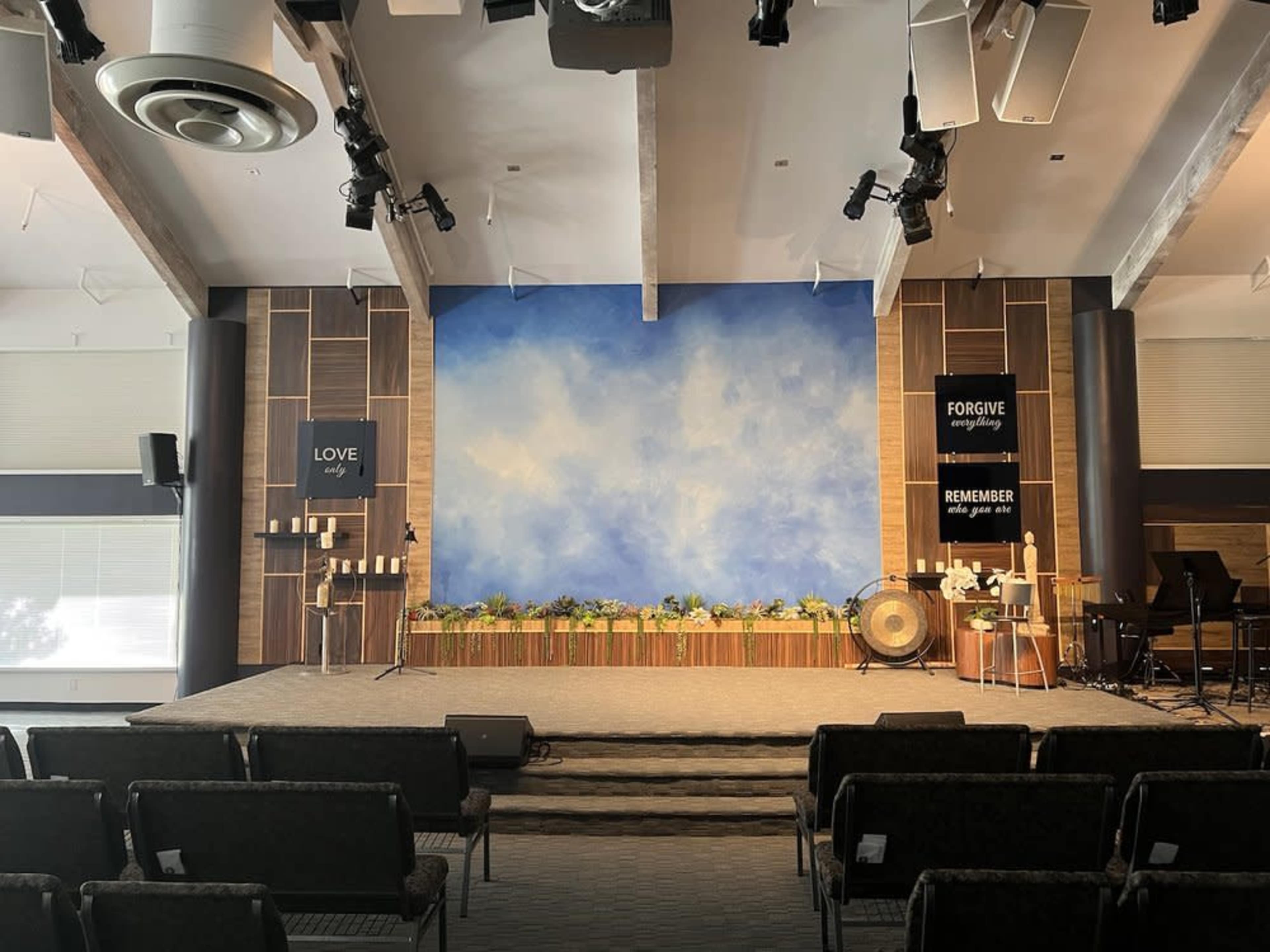 A church stage with a blue sky backdrop, candles, and decorative plants, along with signs that read "LOVE," "FORGIVE," and "REMEMBER."