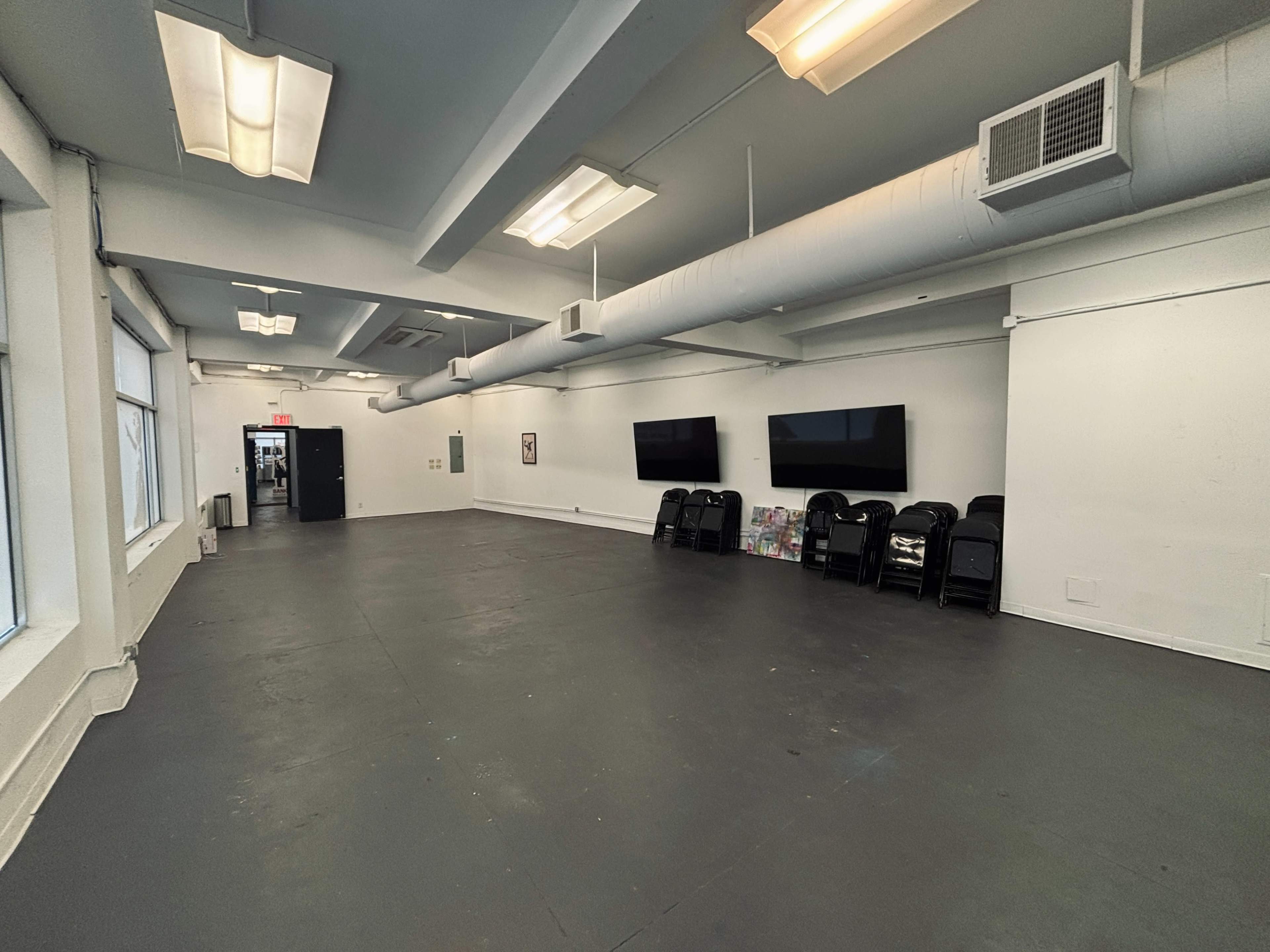 The image shows a spacious, empty room with a gray floor, white walls, and two large television screens mounted on one wall, while several chairs are stacked against another wall.