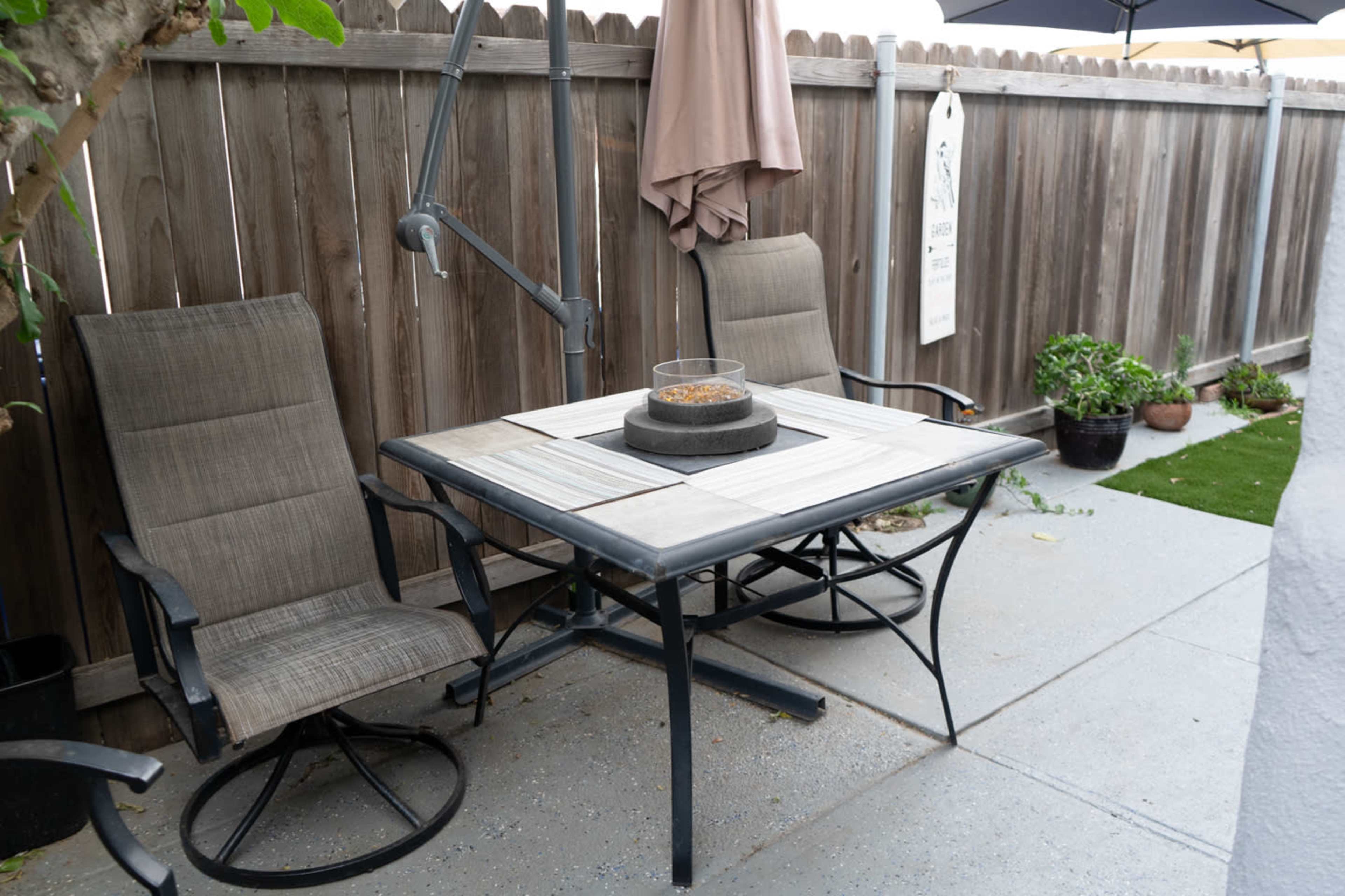 A patio table with two folding chairs and a fire pit is set against a wooden fence.