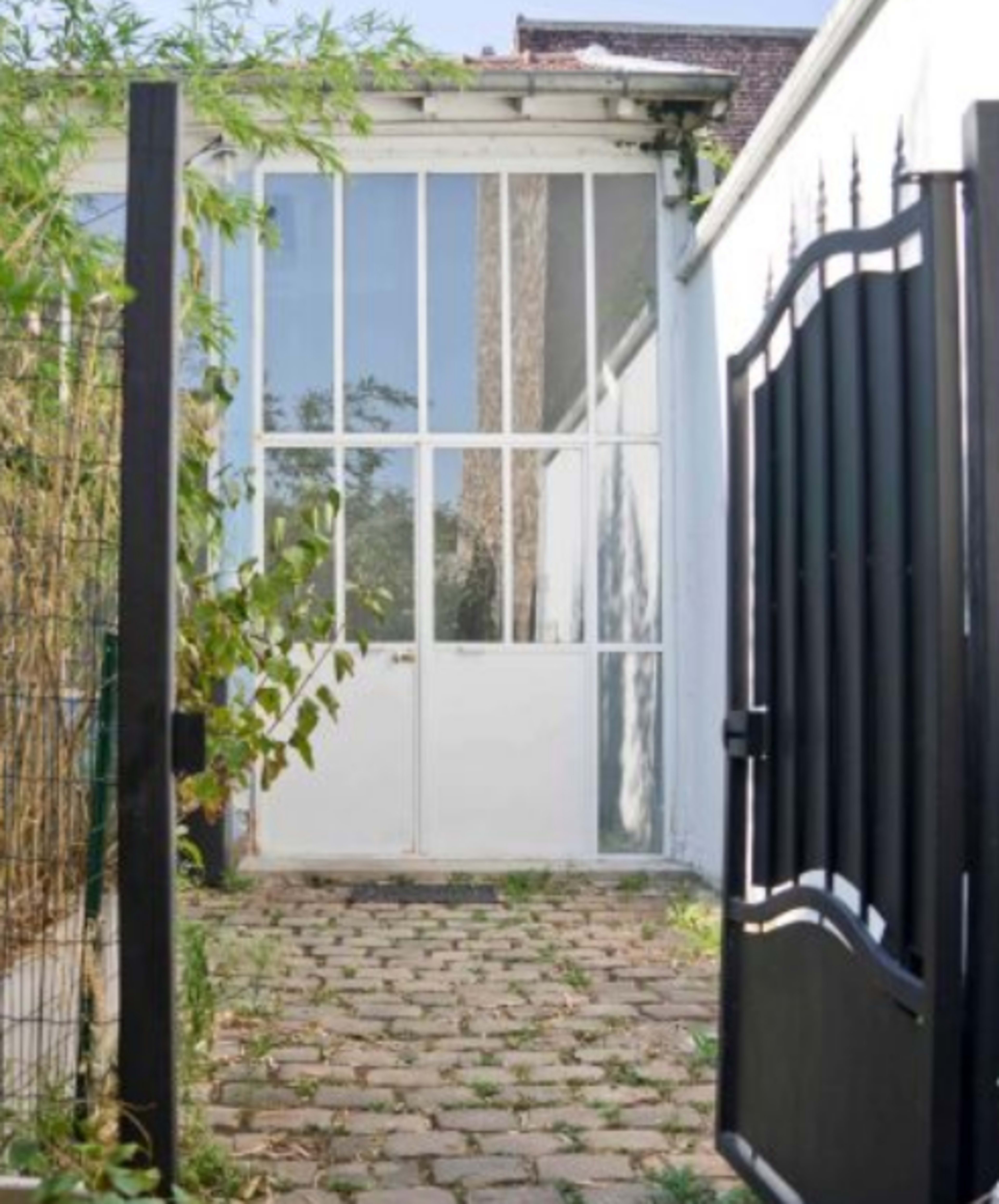 The image shows a black gate opening to a pathway lined with cobblestones leading to a glass-paneled door set against a white wall.