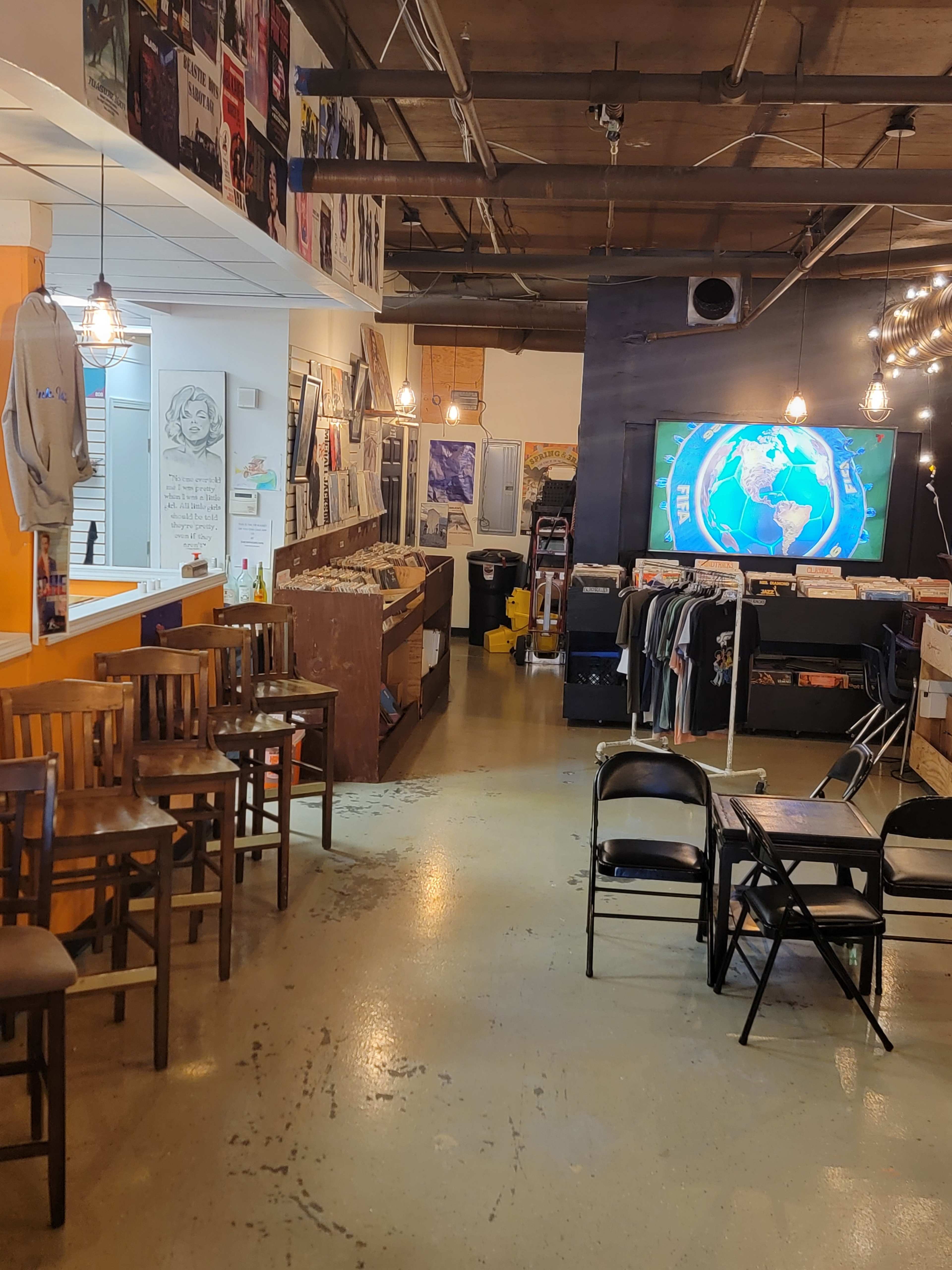 The image shows a record shop interior featuring shelves with vinyl records, display areas for T-shirts, and seating arranged for customers.