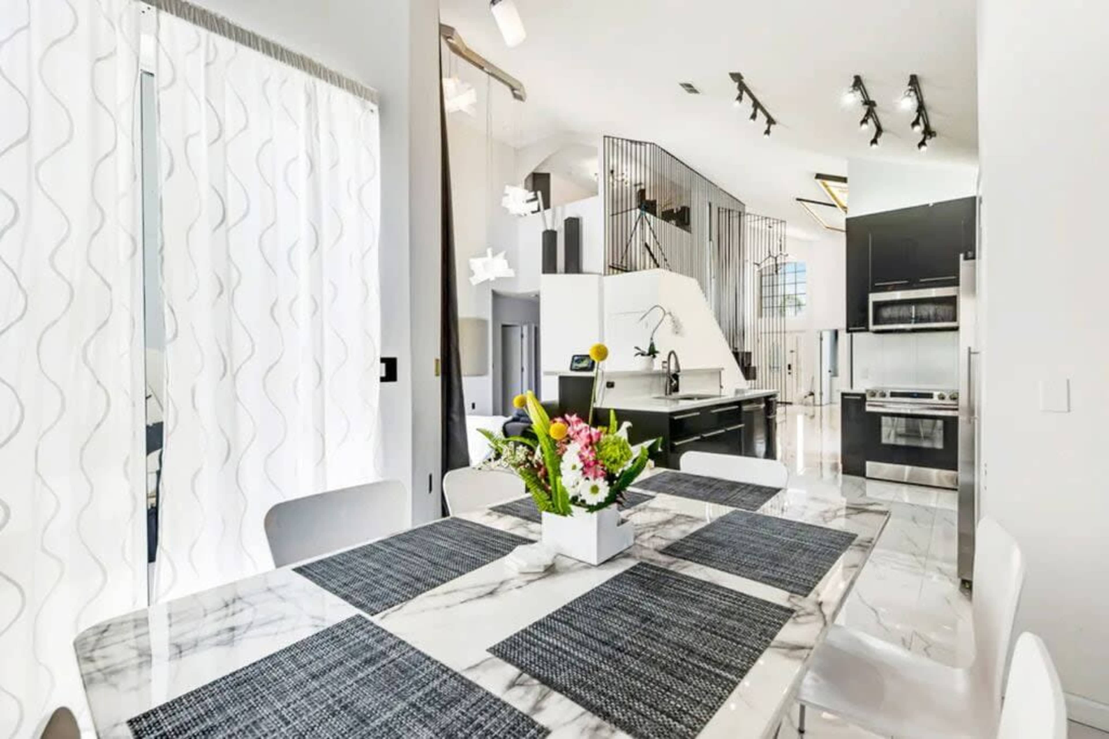 A modern kitchen and dining area feature a marble table with a floral centerpiece, large windows draped with sheer curtains, and sleek cabinetry with stainless steel appliances.
