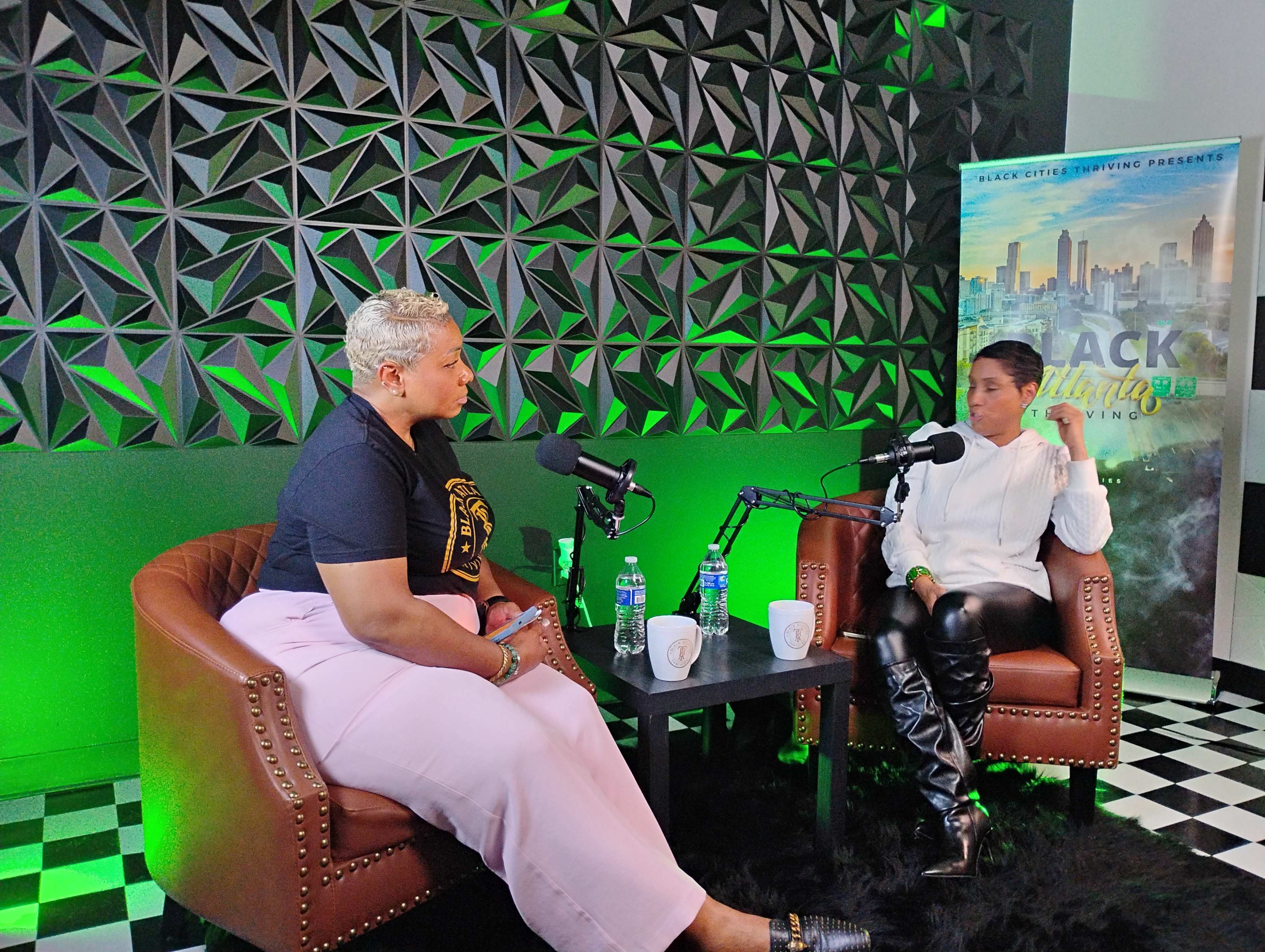 Two women are engaged in a conversation while sitting on brown chairs in a recording studio with green accents and a city skyline banner in the background.