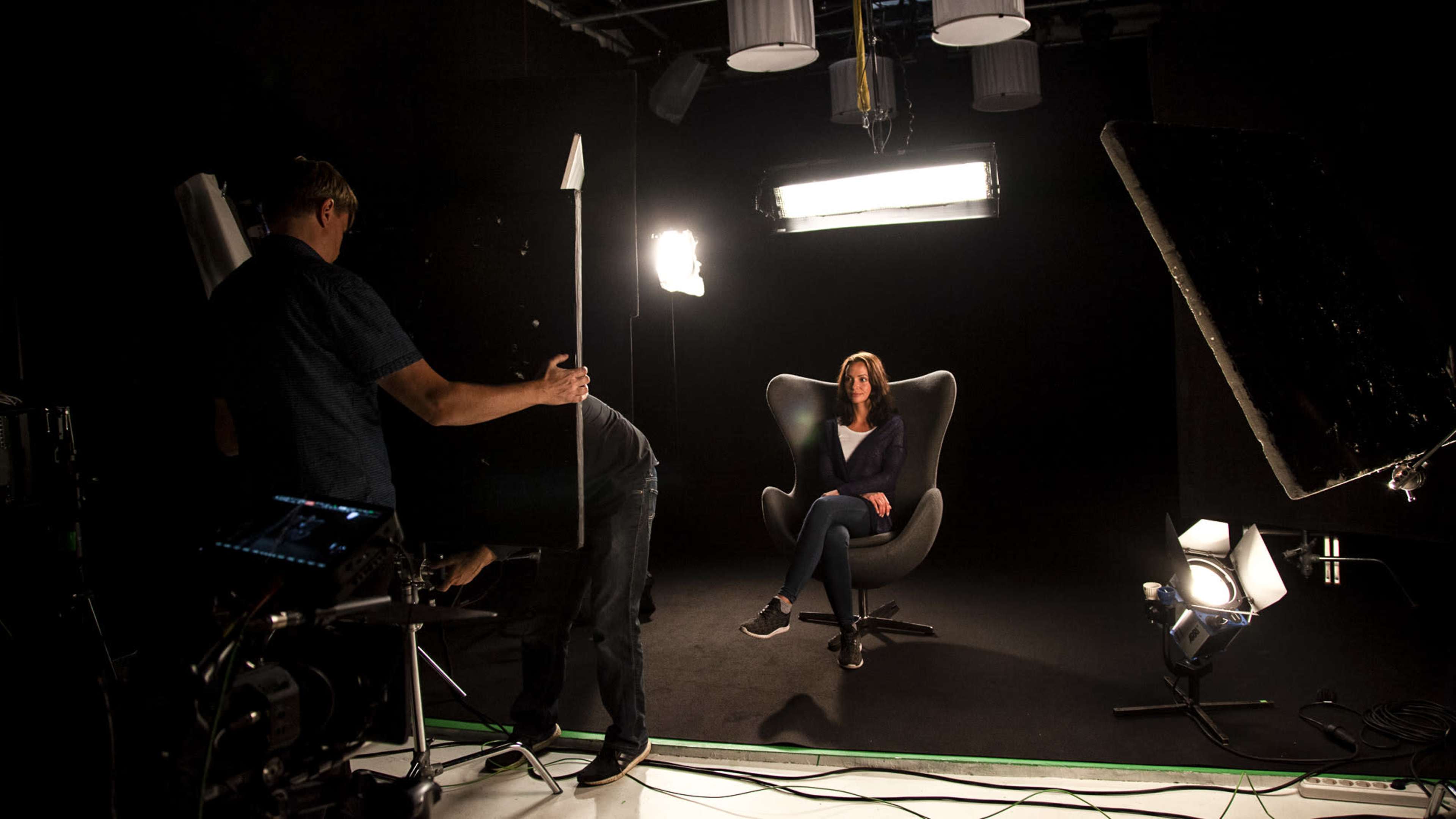 A person sits in a chair while a crew member adjusts lights and equipment in a studio setting.