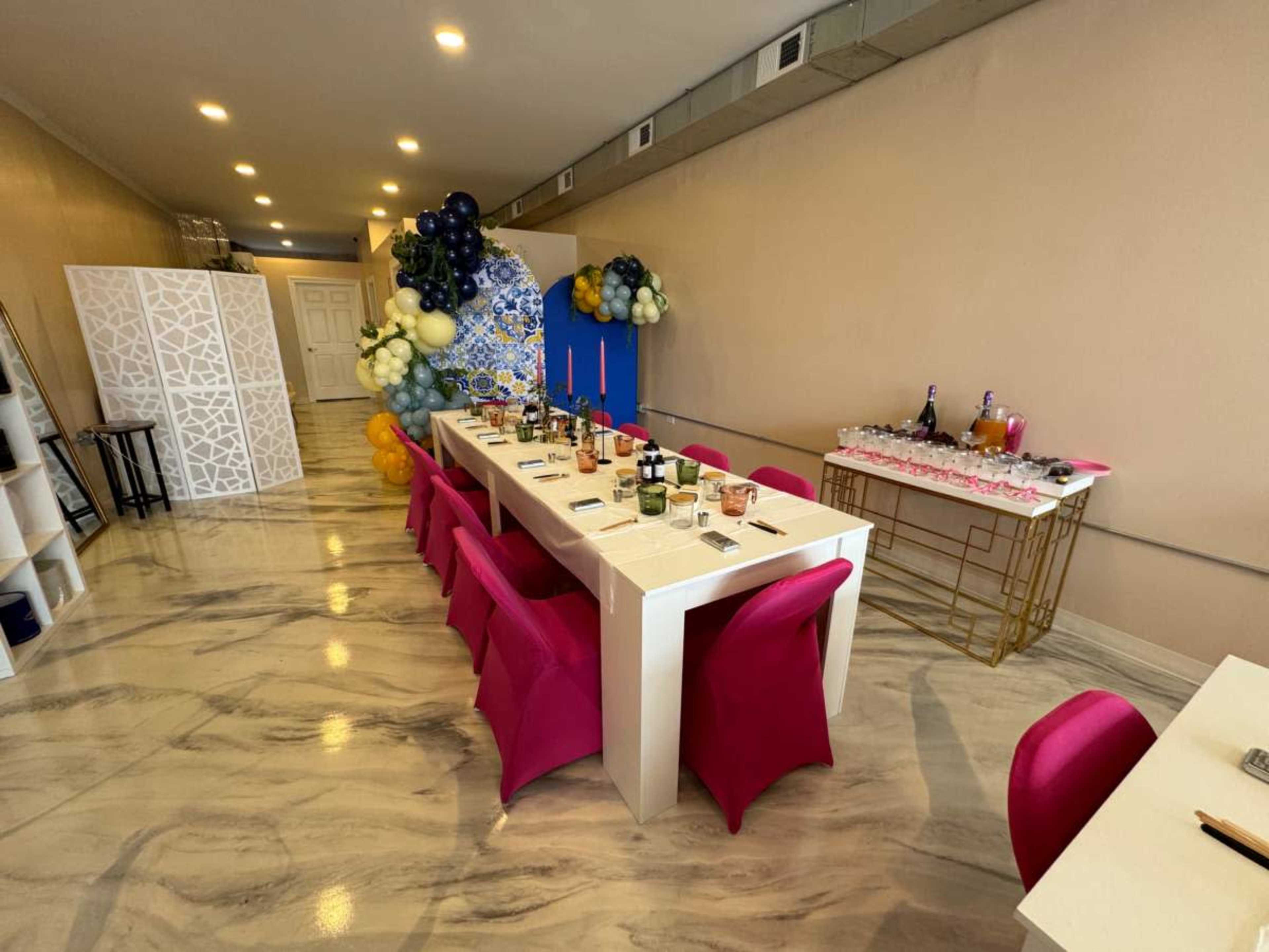 A long dining table is set with various drinks and decorations in a room featuring a colorful balloon arrangement and a side table with additional beverages.