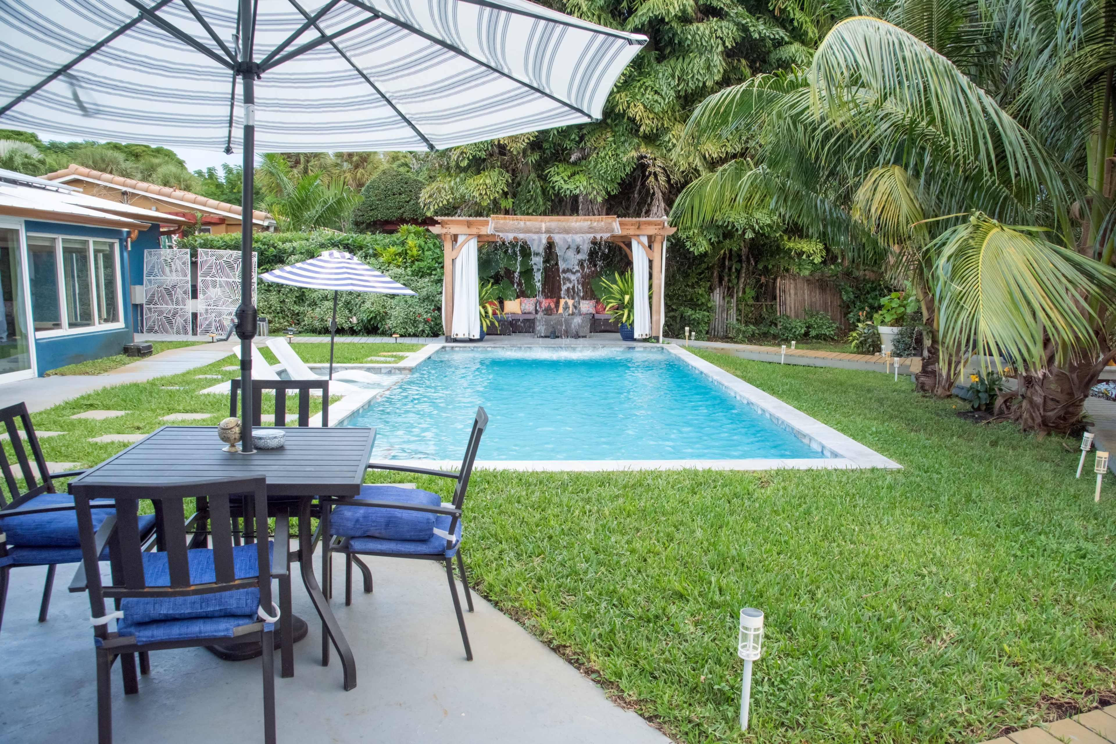 The image shows a backyard with a swimming pool surrounded by a lawn, a patio table and chairs, and a shaded area with a water feature.