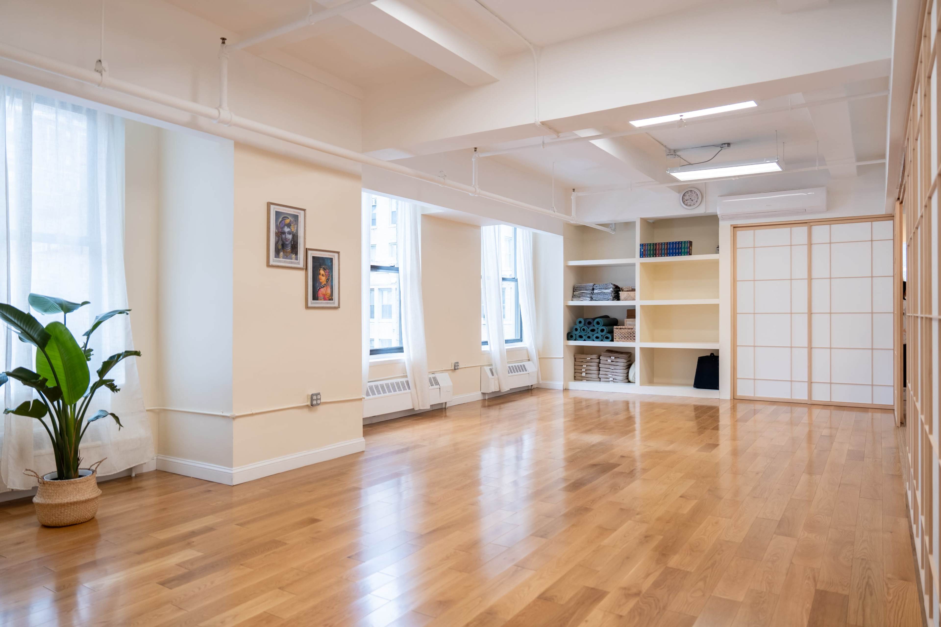The image shows a spacious, well-lit room with wooden flooring, large windows, and minimal furnishings, including a plant and shelves.