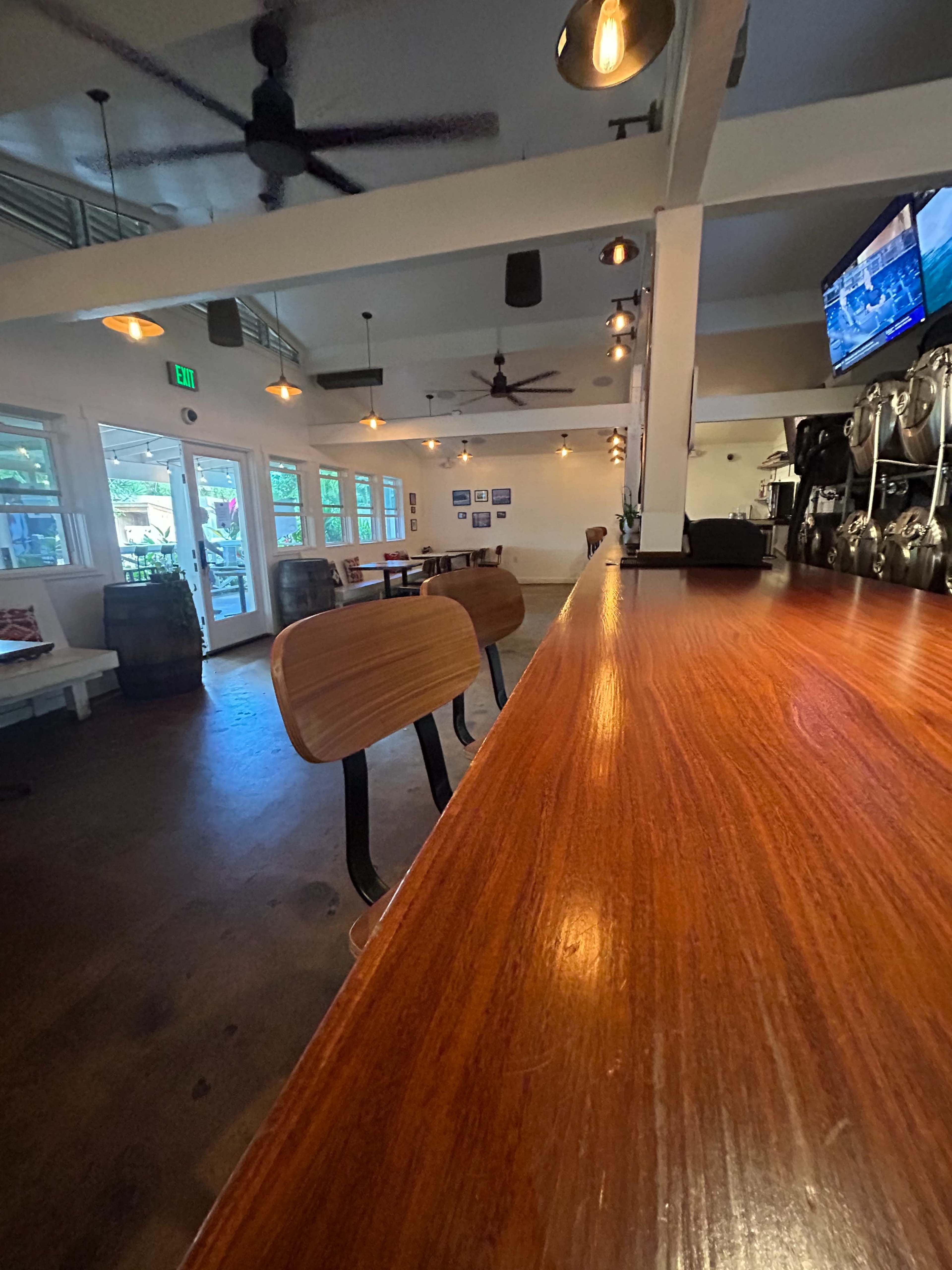 The image shows the interior of a bar or restaurant featuring a polished wooden counter, wooden stools, and ceiling fans, with a television mounted on the wall.