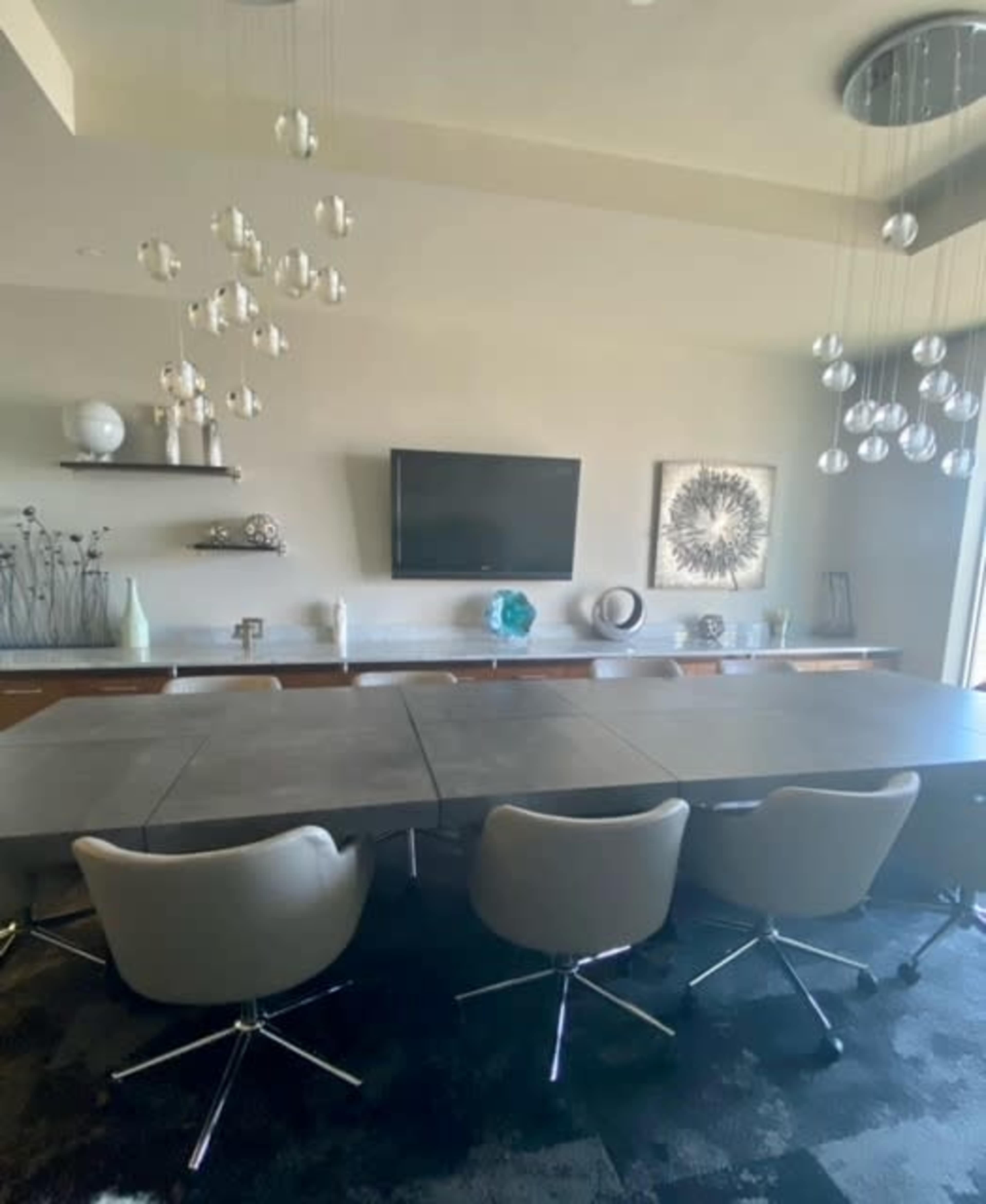 The image shows a modern conference room with a long table, several chrome-accented chairs, a wall-mounted television, and decorative items on shelves.