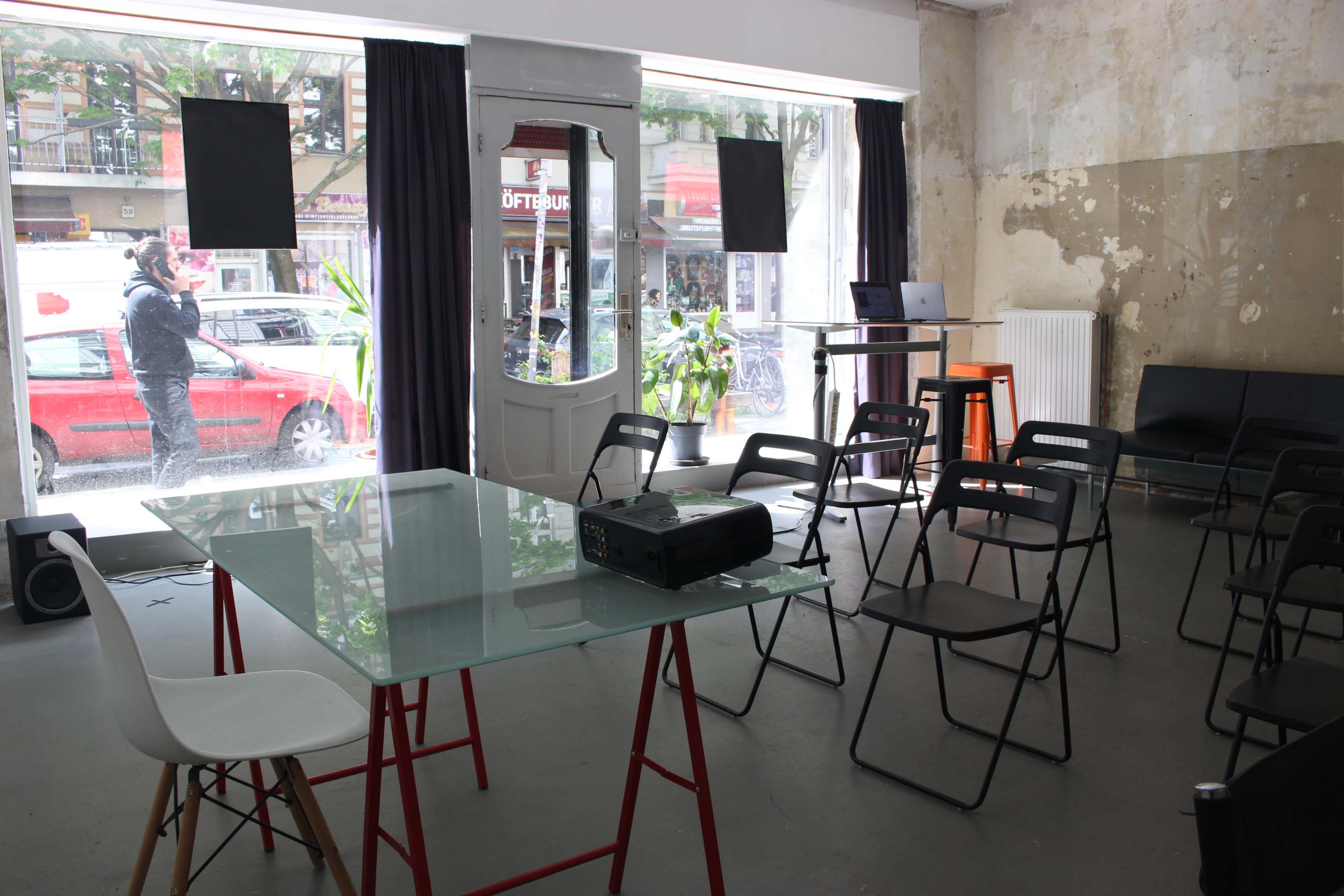 The image shows an indoor setting with a glass table and black folding chairs arranged for a meeting, alongside a projector and a view of a street outside.