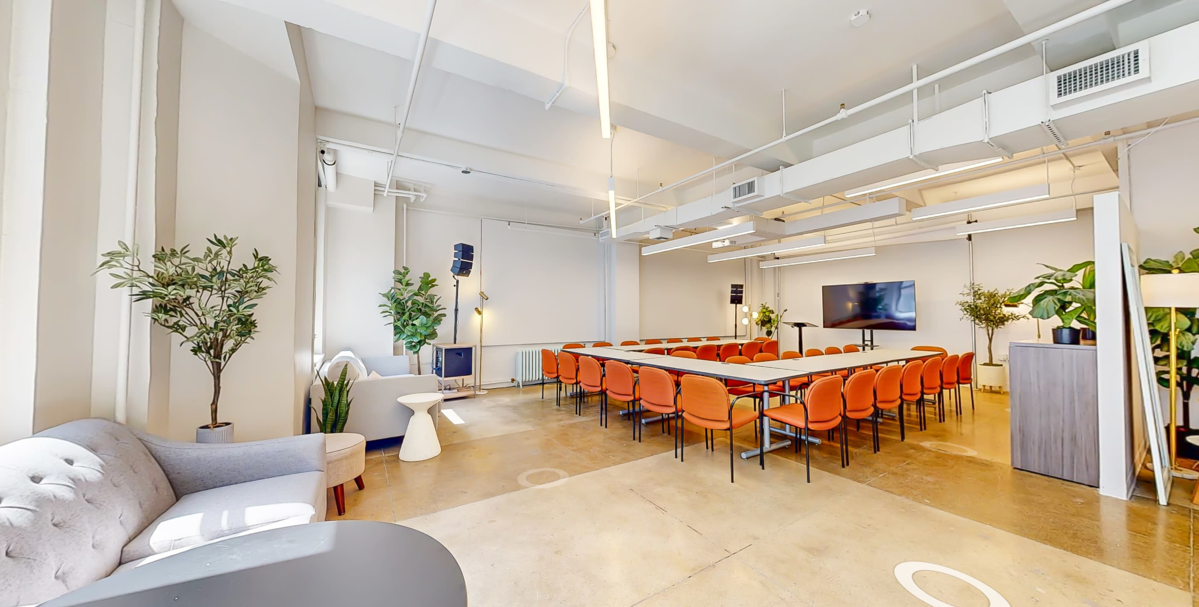 A modern conference room with a long table surrounded by orange chairs, potted plants, and a large screen at one end.