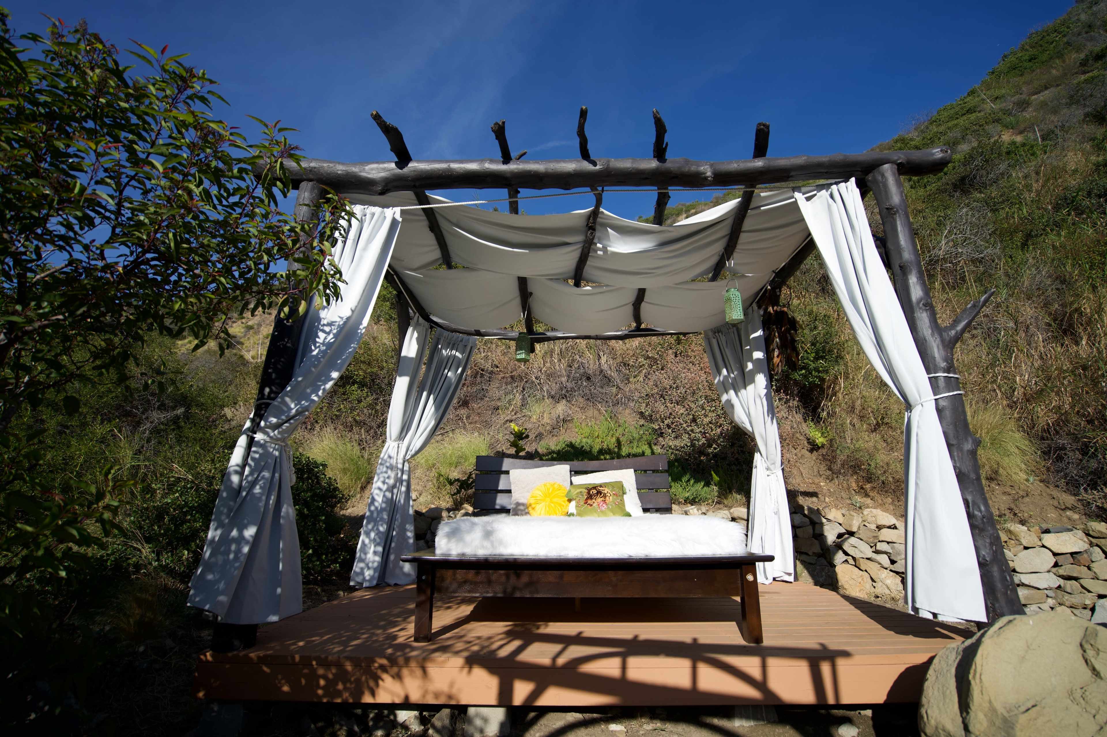 A wooden gazebo with white drapes and a mattress sits on a wooden deck surrounded by lush greenery and hills.