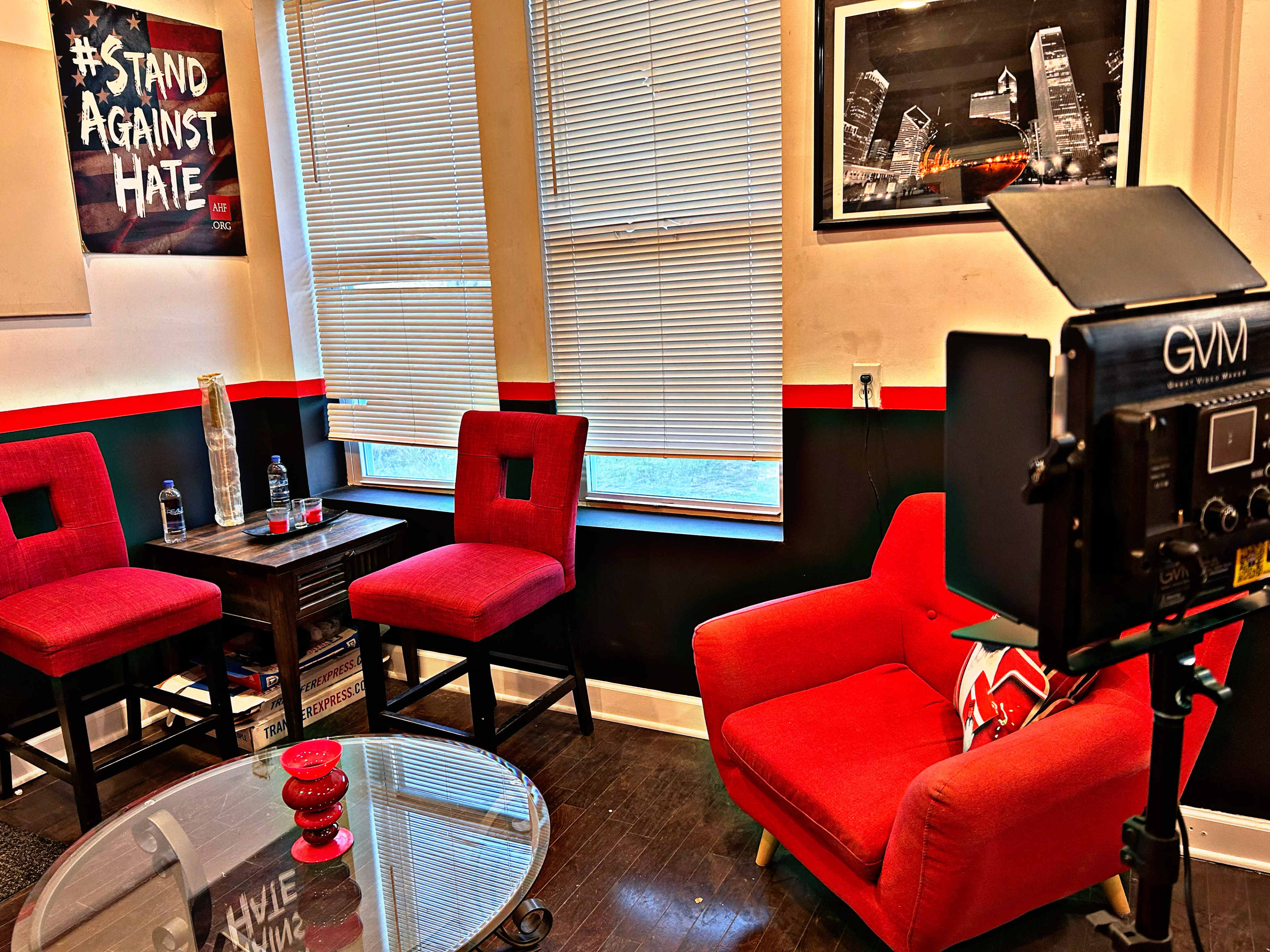 The image shows a cozy interior space with two red chairs, a small table, and a camera setup facing a wall adorned with a "Stand Against Hate" poster and a cityscape photo.