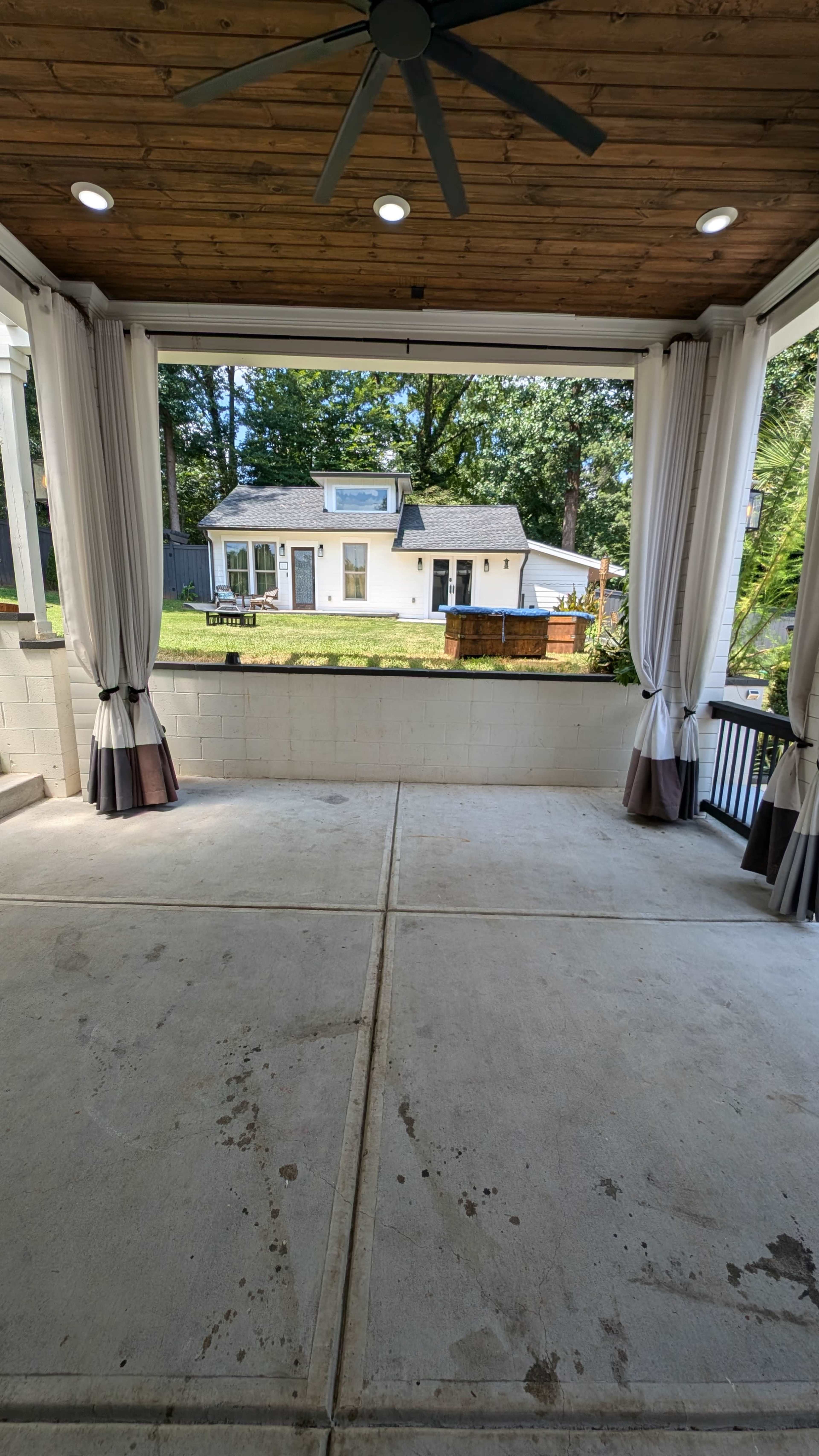 A covered porch with concrete flooring, featuring white curtains and a view of a house and lawn in the background.