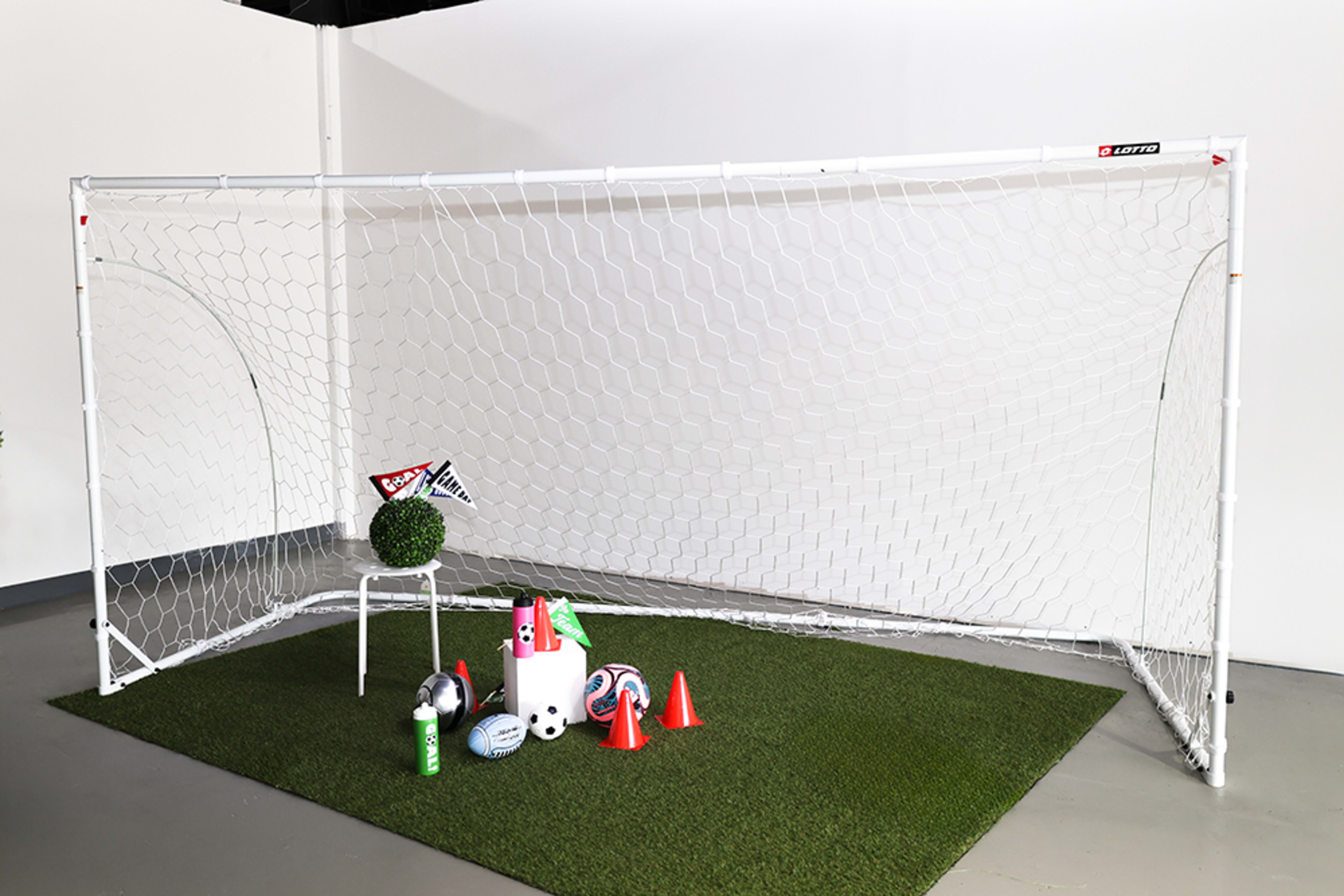 The image shows a soccer goal on a grassy surface with various sports equipment, including cones, balls, and colorful flags arranged nearby.
