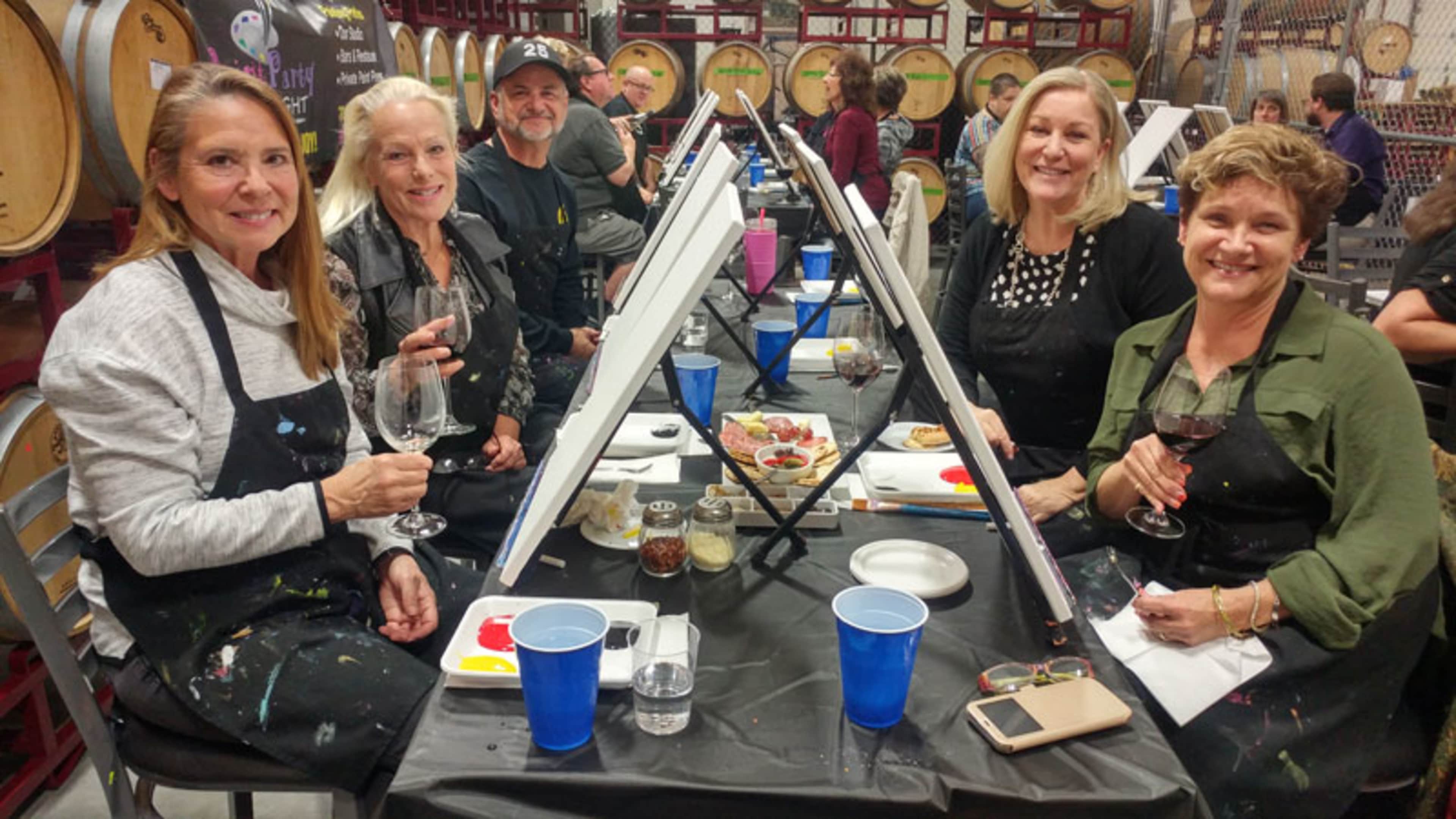 A group of five women sits at a table with easels in a wine tasting room, holding wine glasses and surrounded by art supplies and snacks.