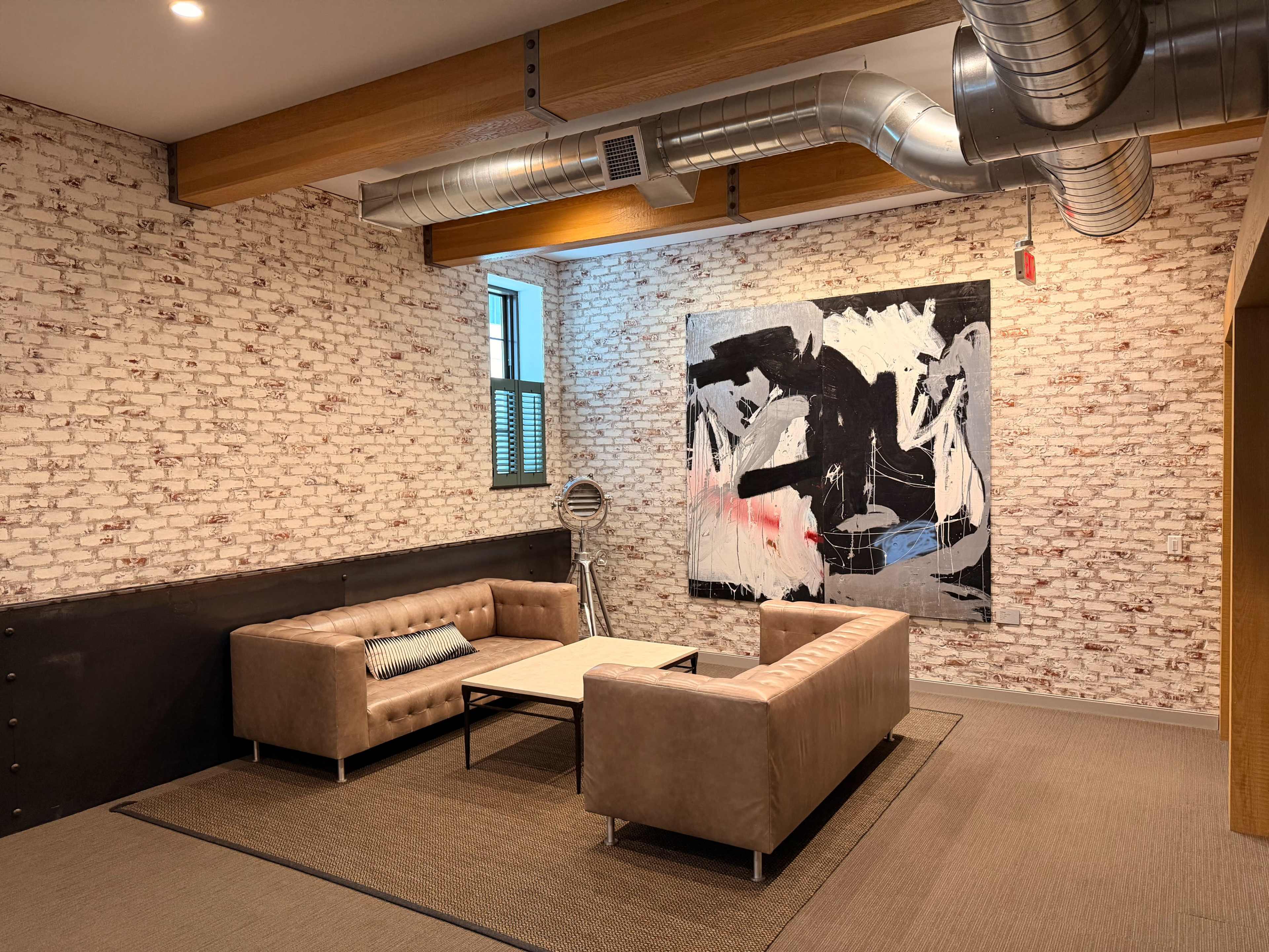 The image shows a modern lounge area with two beige leather sofas, a coffee table, and an abstract painting on the brick wall.