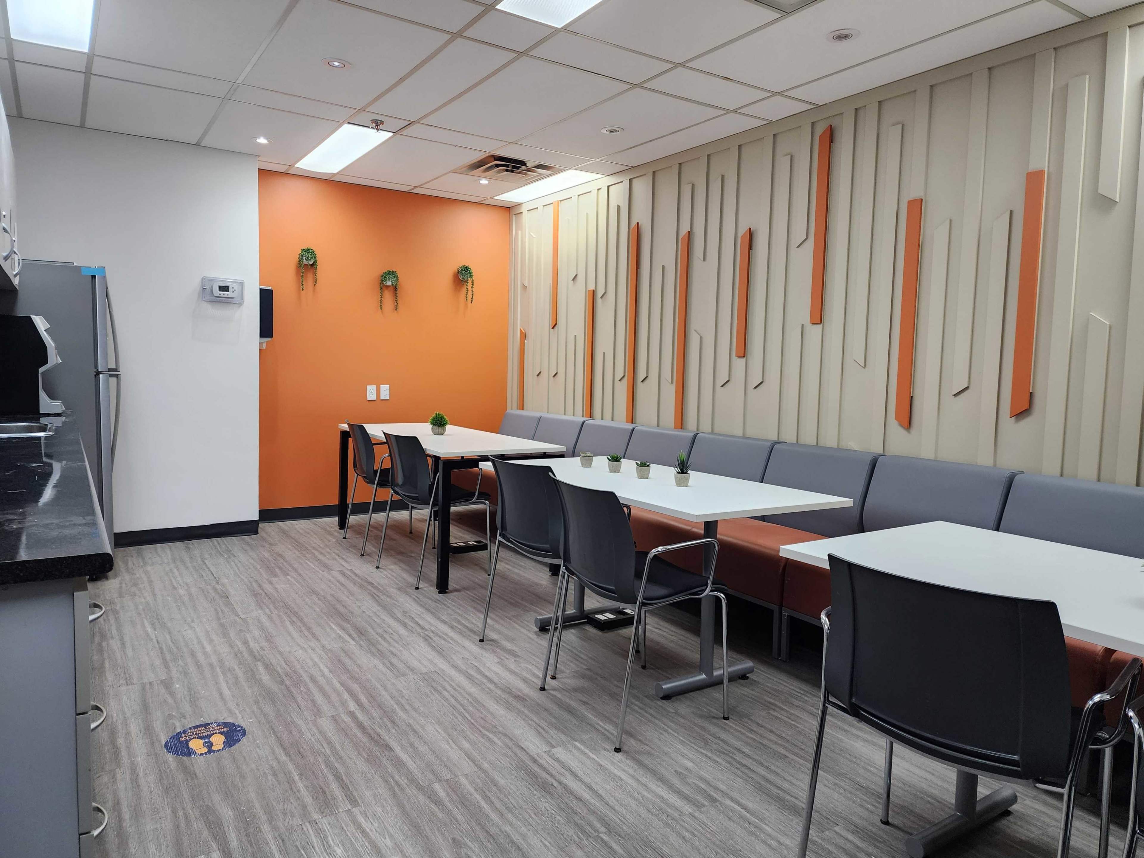 The image shows a modern break room with a gray and orange color scheme, featuring a row of tables and chairs along one side, a kitchenette on the left, and decorative wall elements.