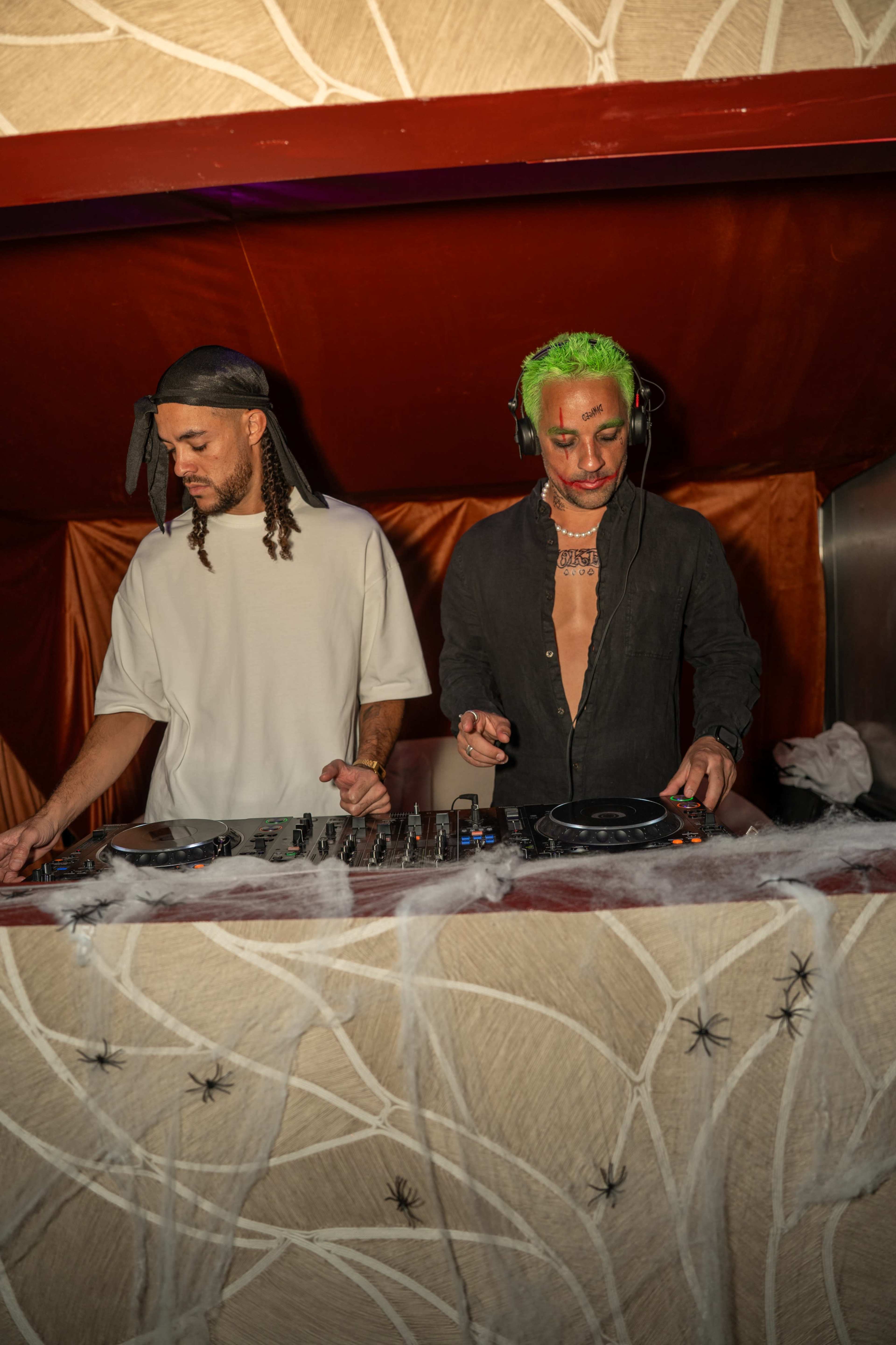 Two DJs are mixing music at a nightclub, with one wearing a white shirt and the other in a black outfit, both positioned behind a decorated DJ booth.