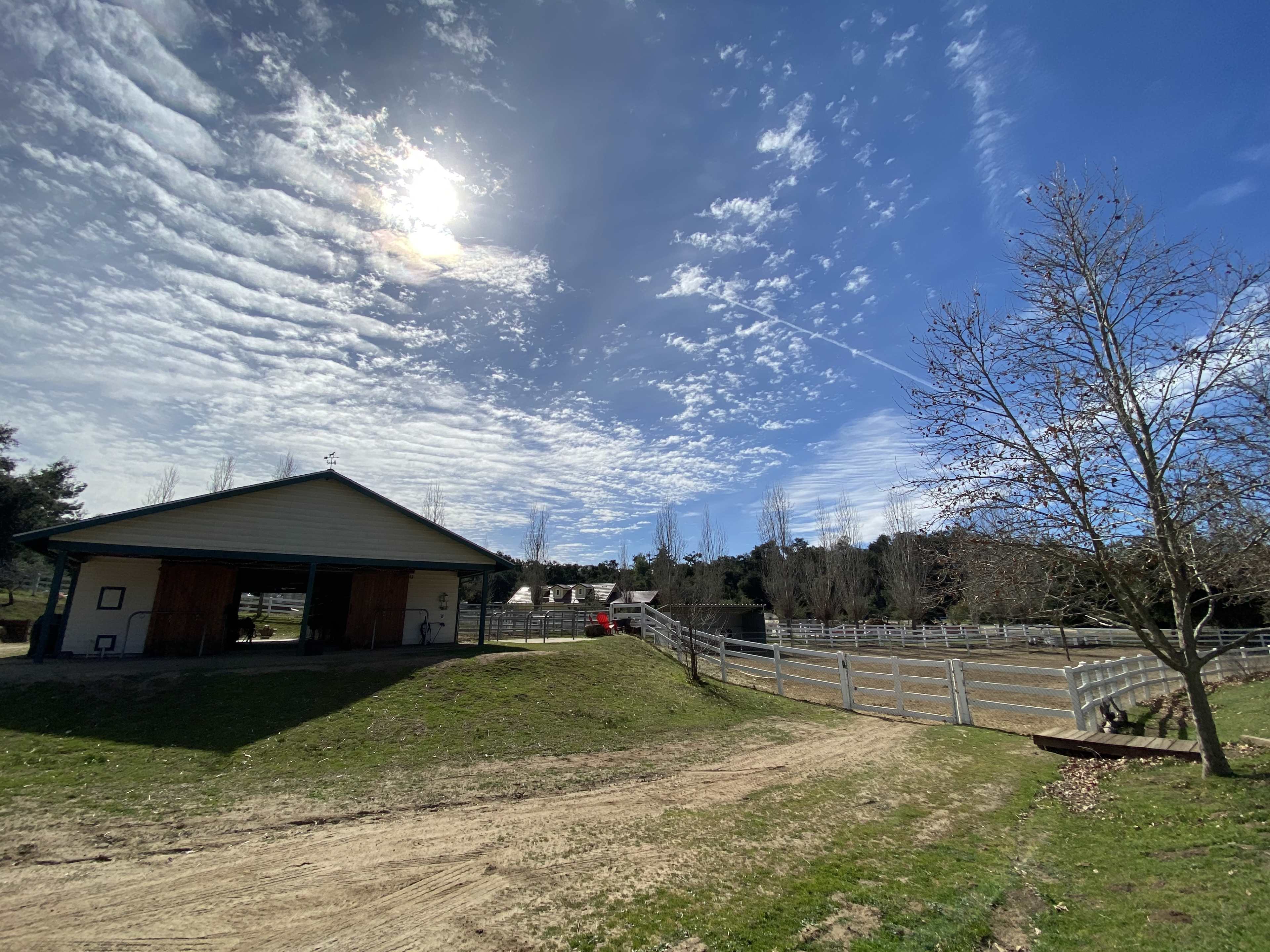 A barn sits beside a fenced horse paddock under a partly cloudy sky.
