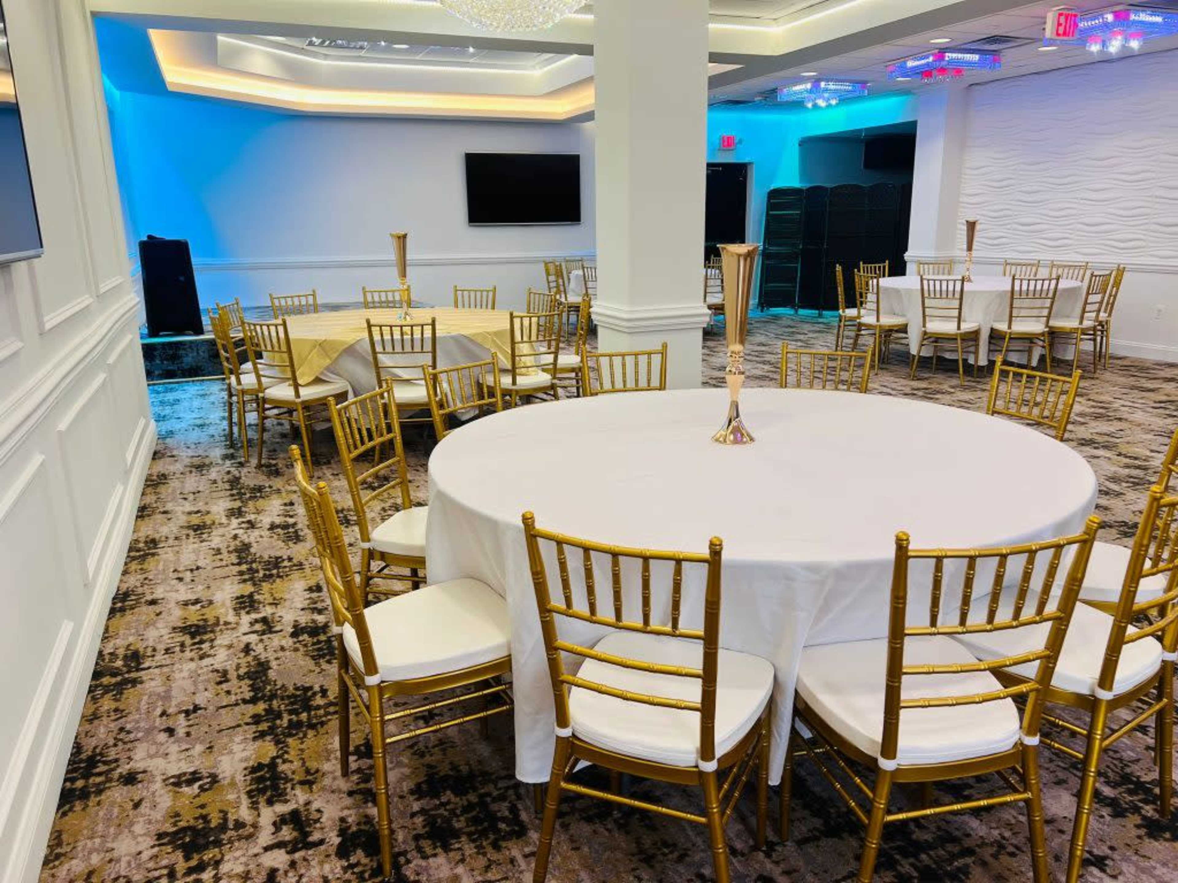 The image shows a banquet room featuring several round tables set with white tablecloths and gold chairs, with a modern, well-lit atmosphere.