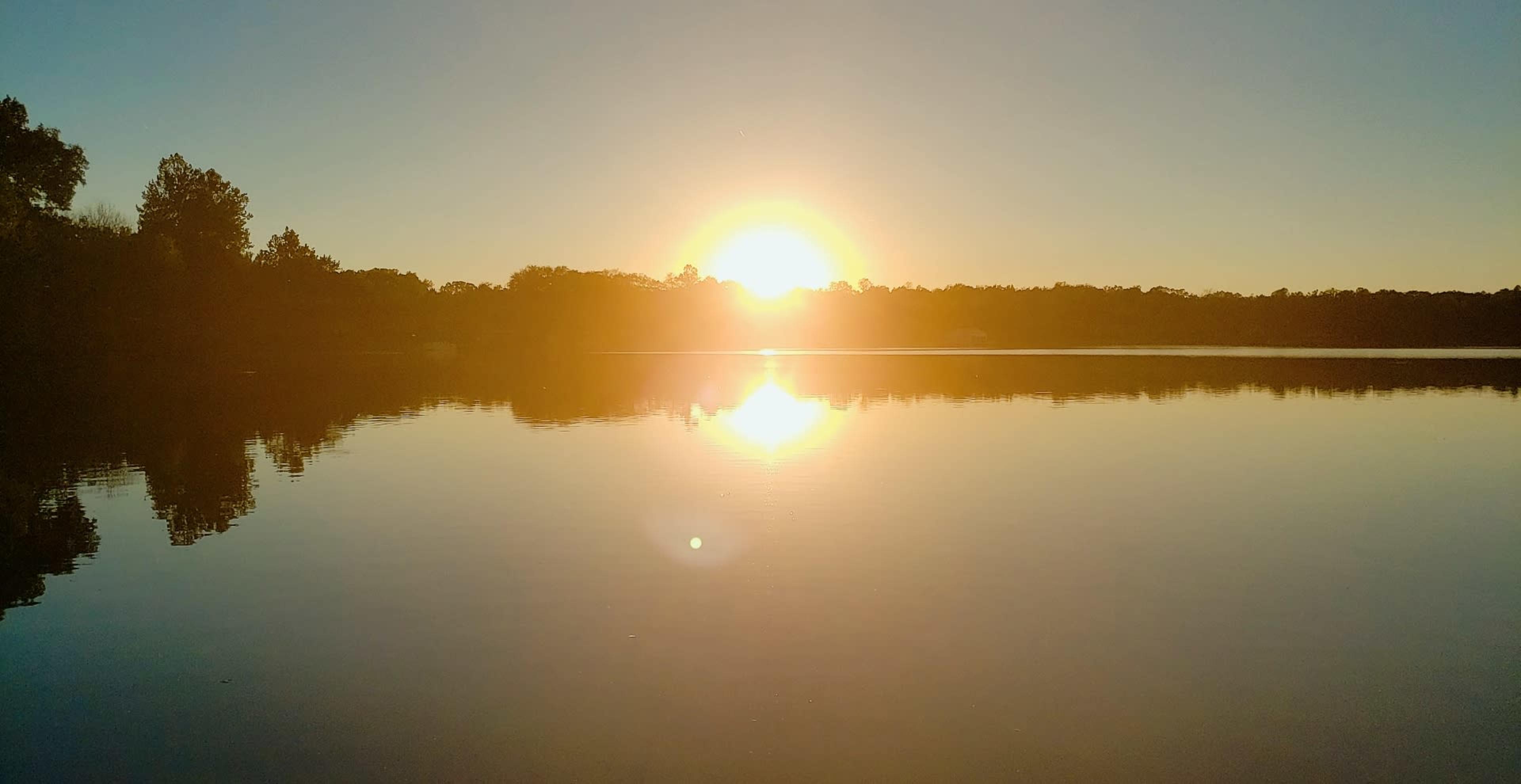 The sun rises over a calm lake, reflecting light on the water's surface.