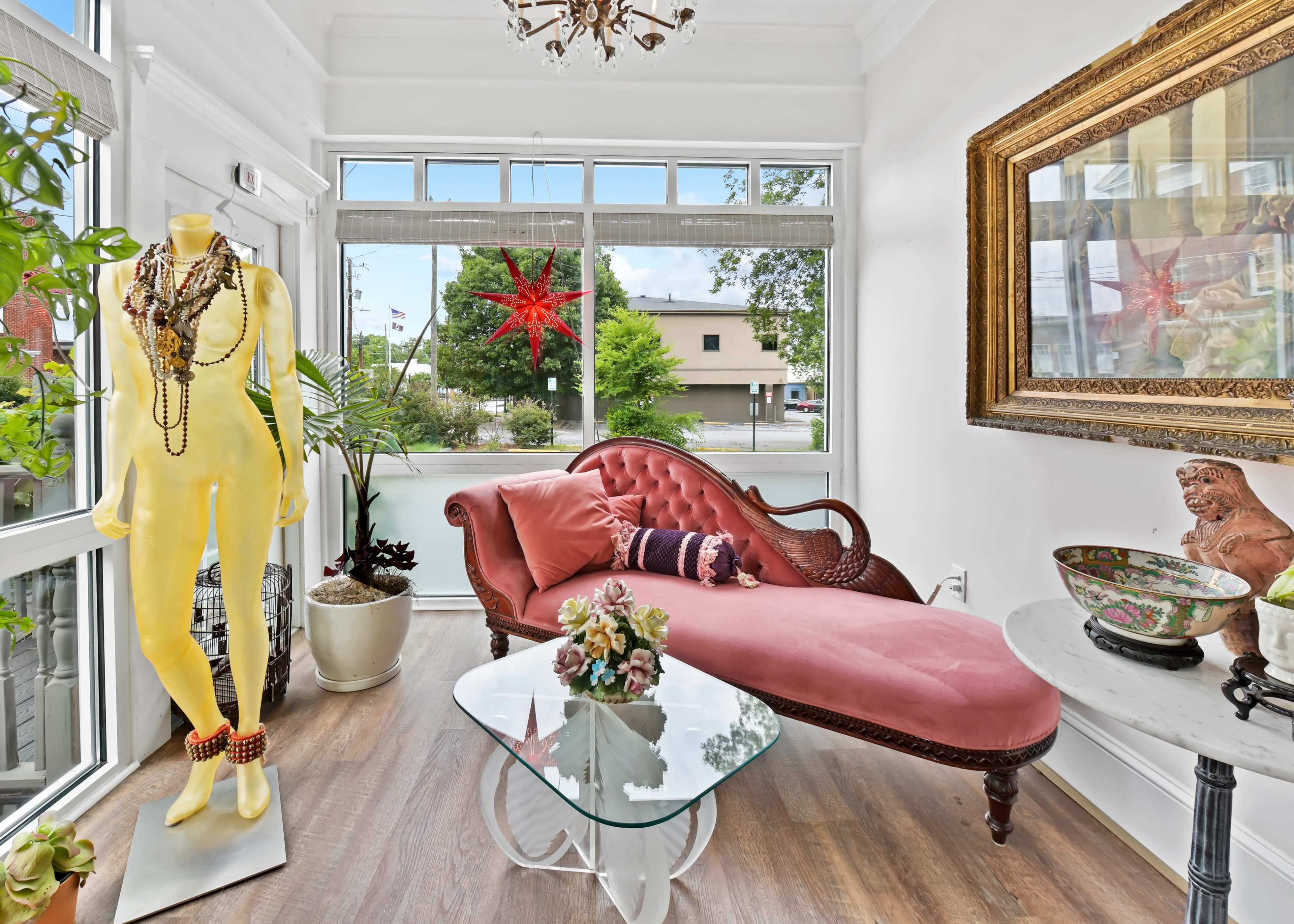 The scene features a bright interior with a vintage chaise lounge, a standing mannequin adorned with accessories, a glass coffee table, and decorative plants near large windows.